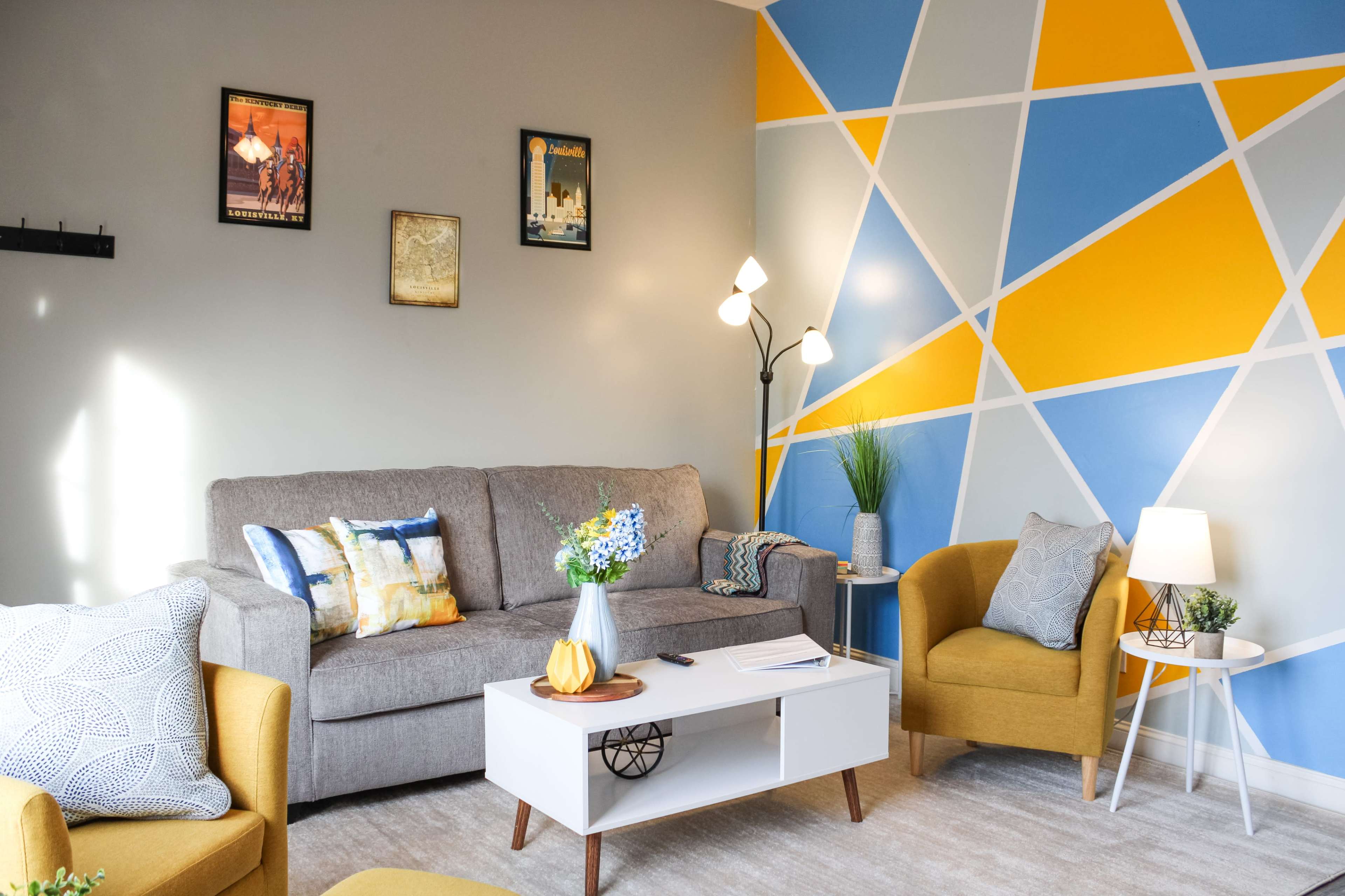 A modern living room with a gray sofa, two yellow chairs, a coffee table, and a colorful geometric wall design in blue and yellow.