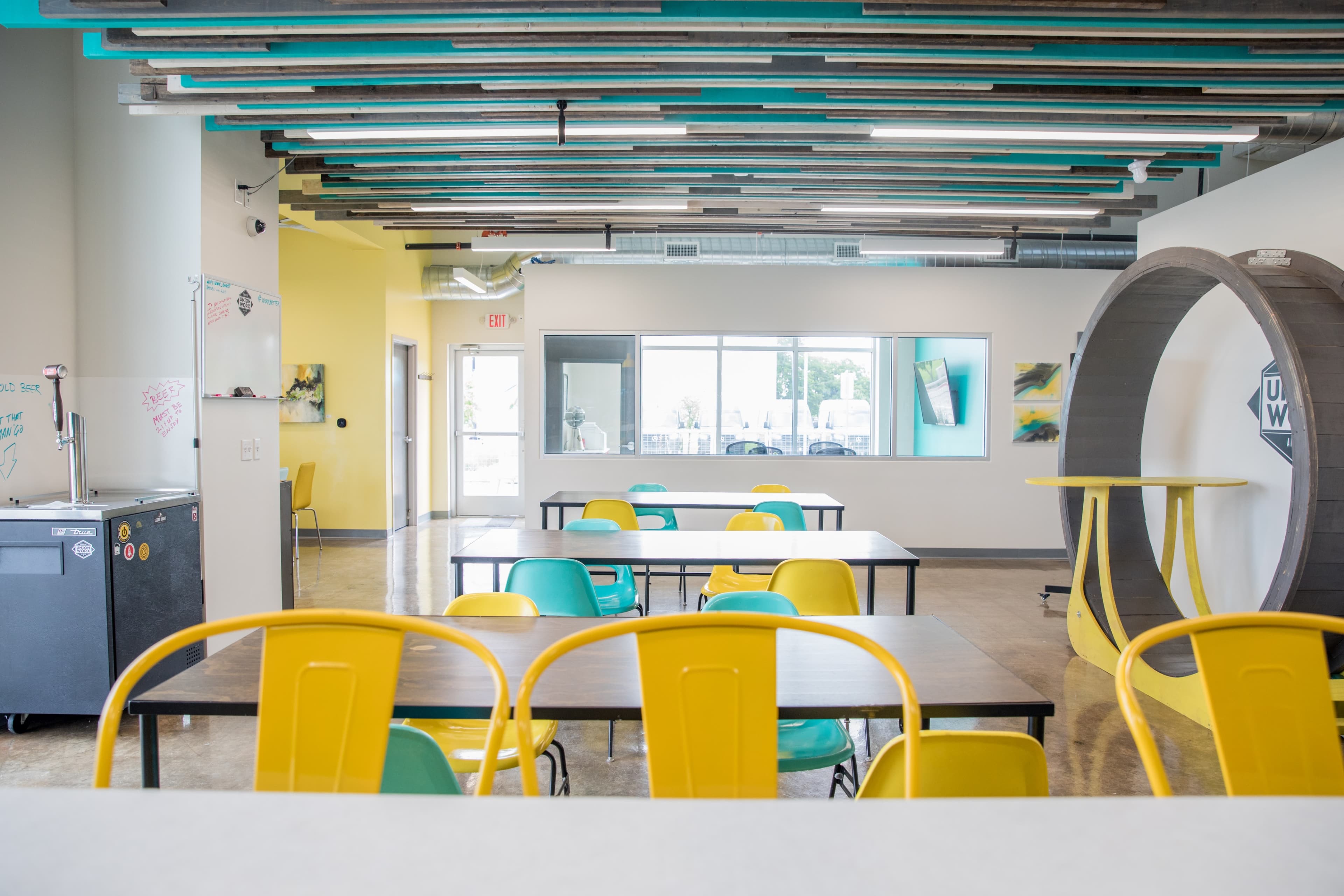 A modern, open communal space with yellow and teal chairs around tables, featuring a unique ceiling design and large windows.