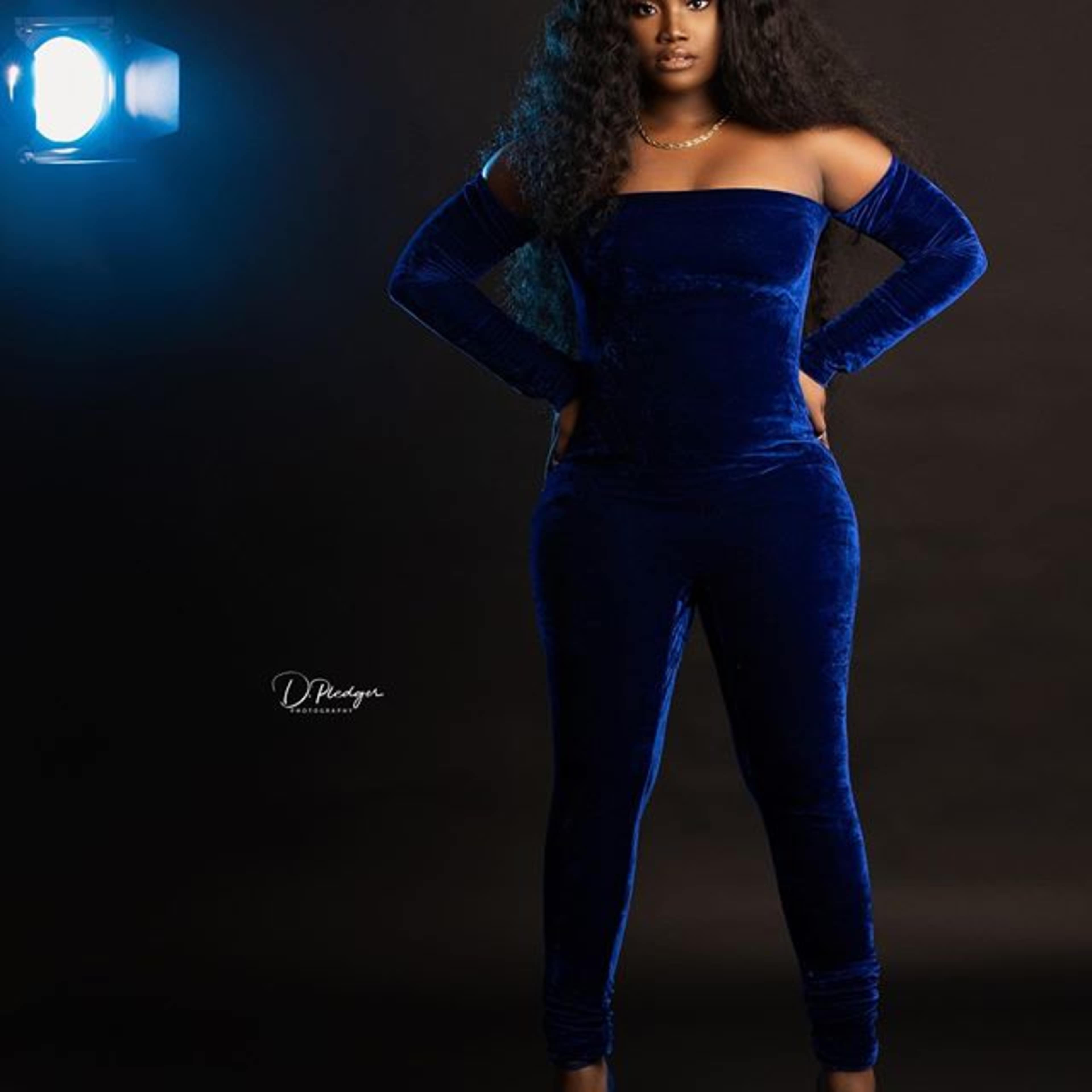 A woman in a blue velvet jumpsuit stands confidently with her hands on her hips in front of a spotlight.