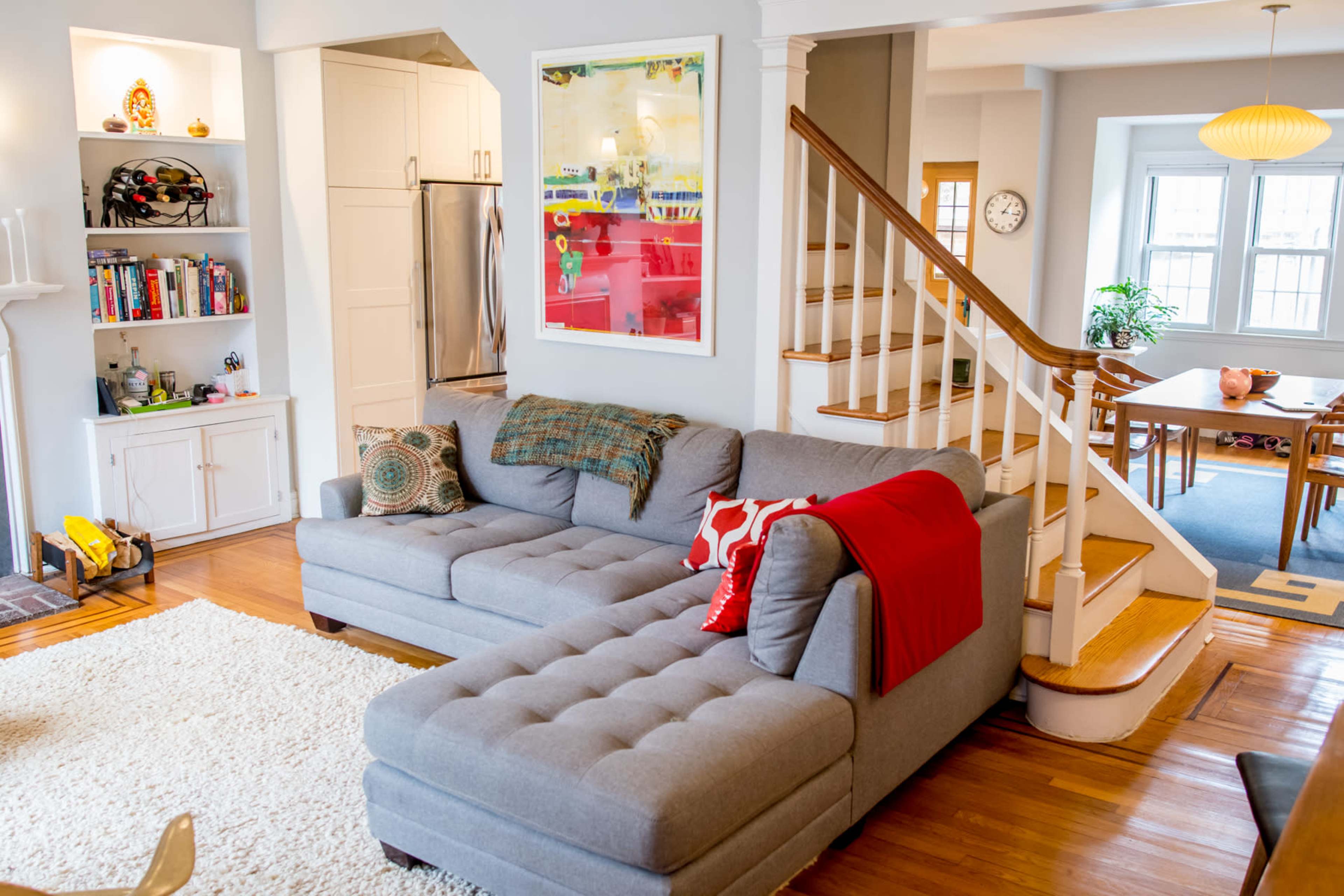 A cozy living room with a gray sectional sofa, a red throw blanket, a wooden staircase, and a dining area visible in the background.
