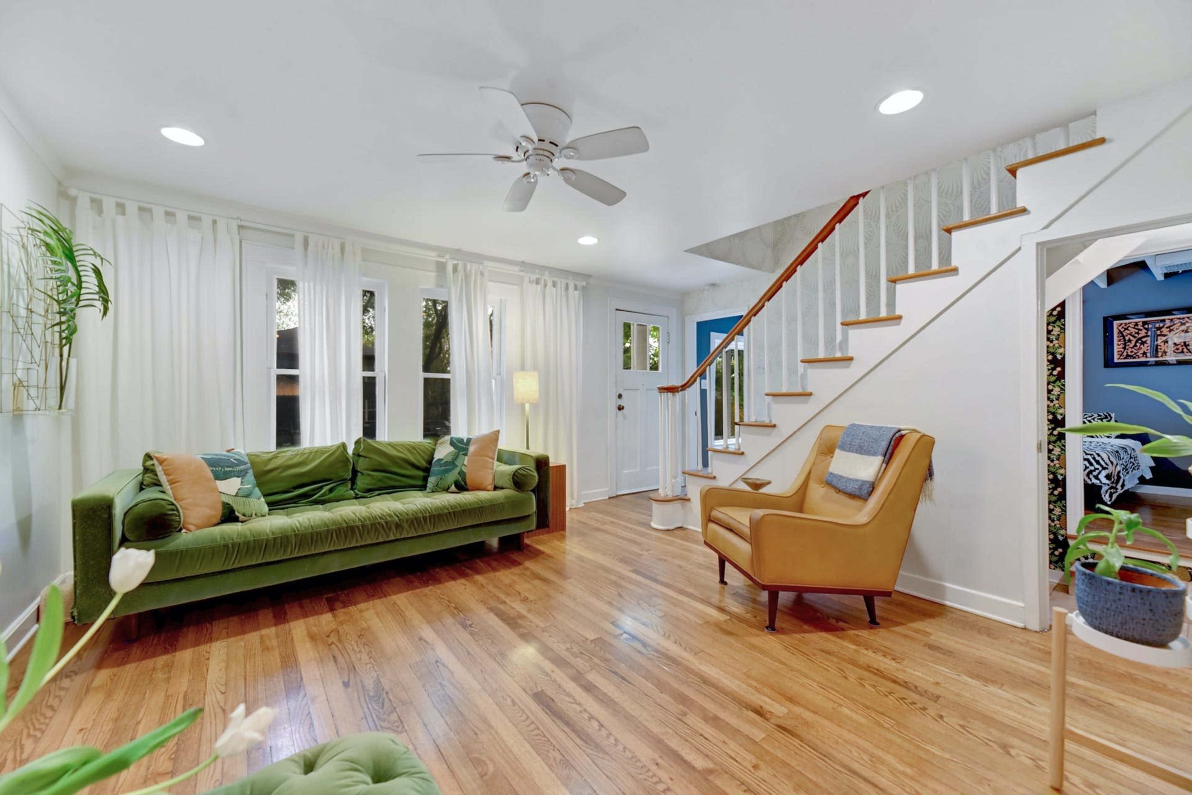 The image shows a bright living room with a green sofa, an armchair, a wooden staircase, and large windows draped with white curtains.