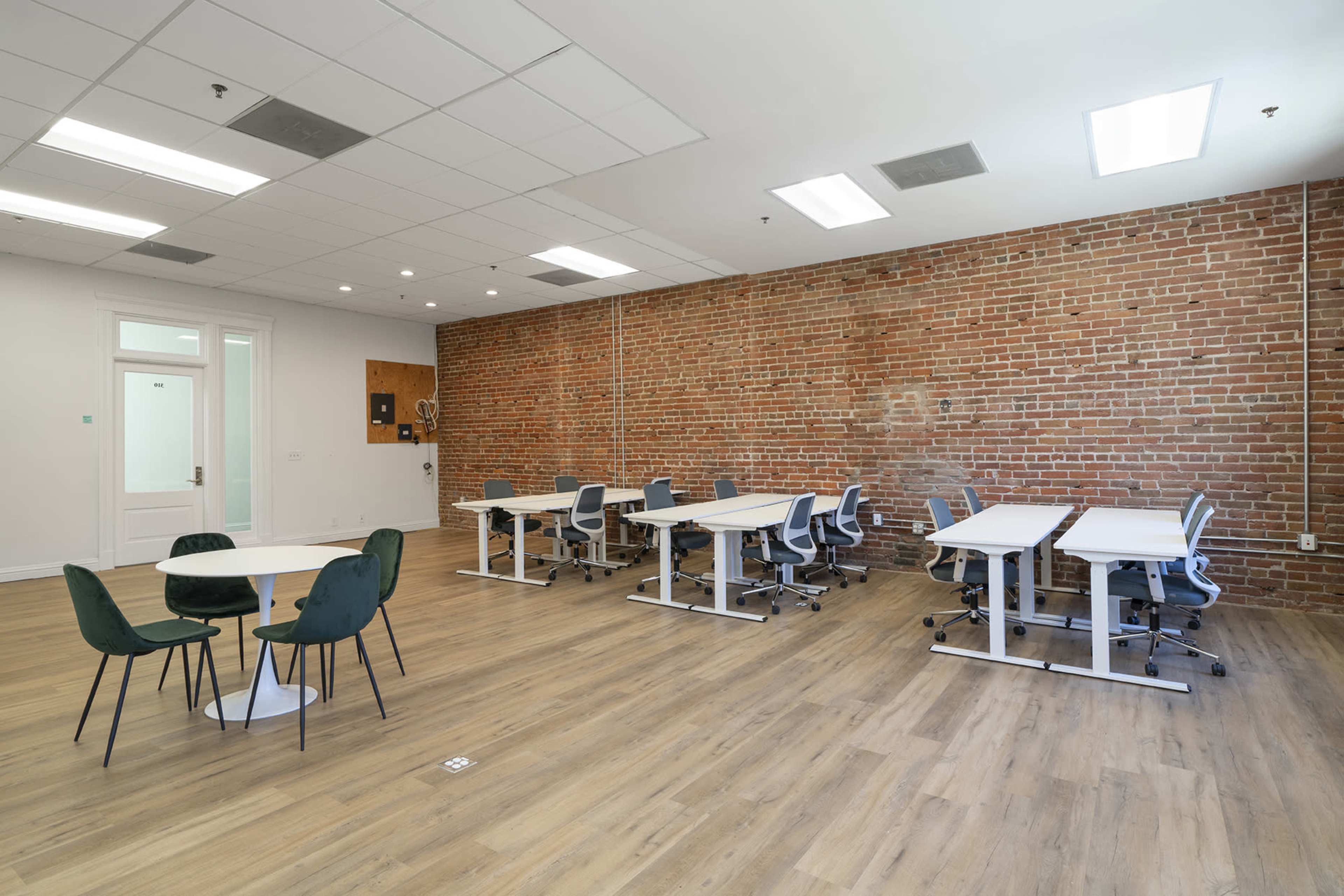 A spacious room with a brick wall, featuring multiple desks and chairs arranged for work, along with a round table and chairs in the center.