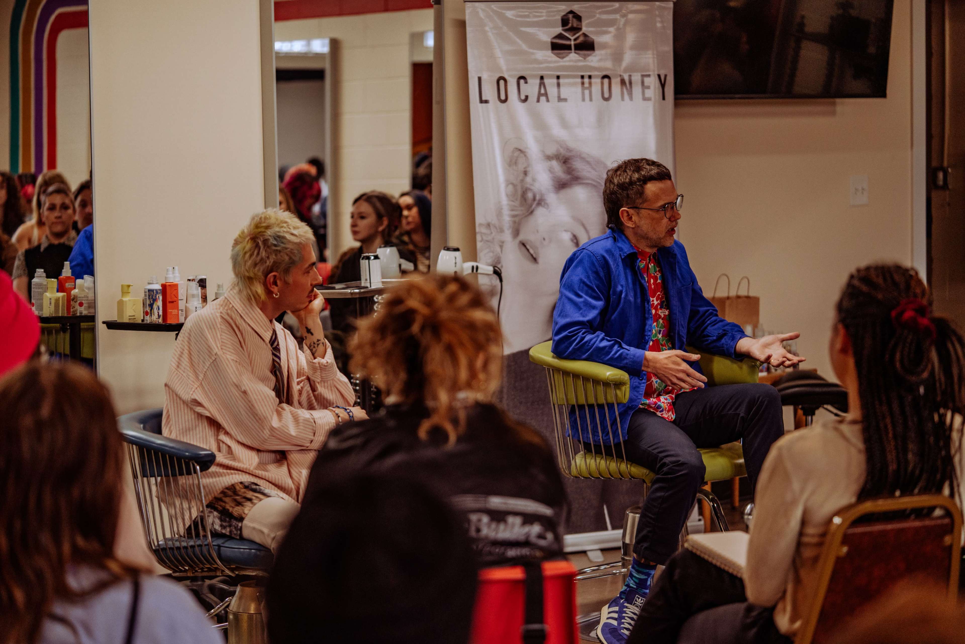 A group of people is seated in a room, listening to a man in glasses and a colorful shirt speaking in front of a banner that reads "LOCAL HONEY."