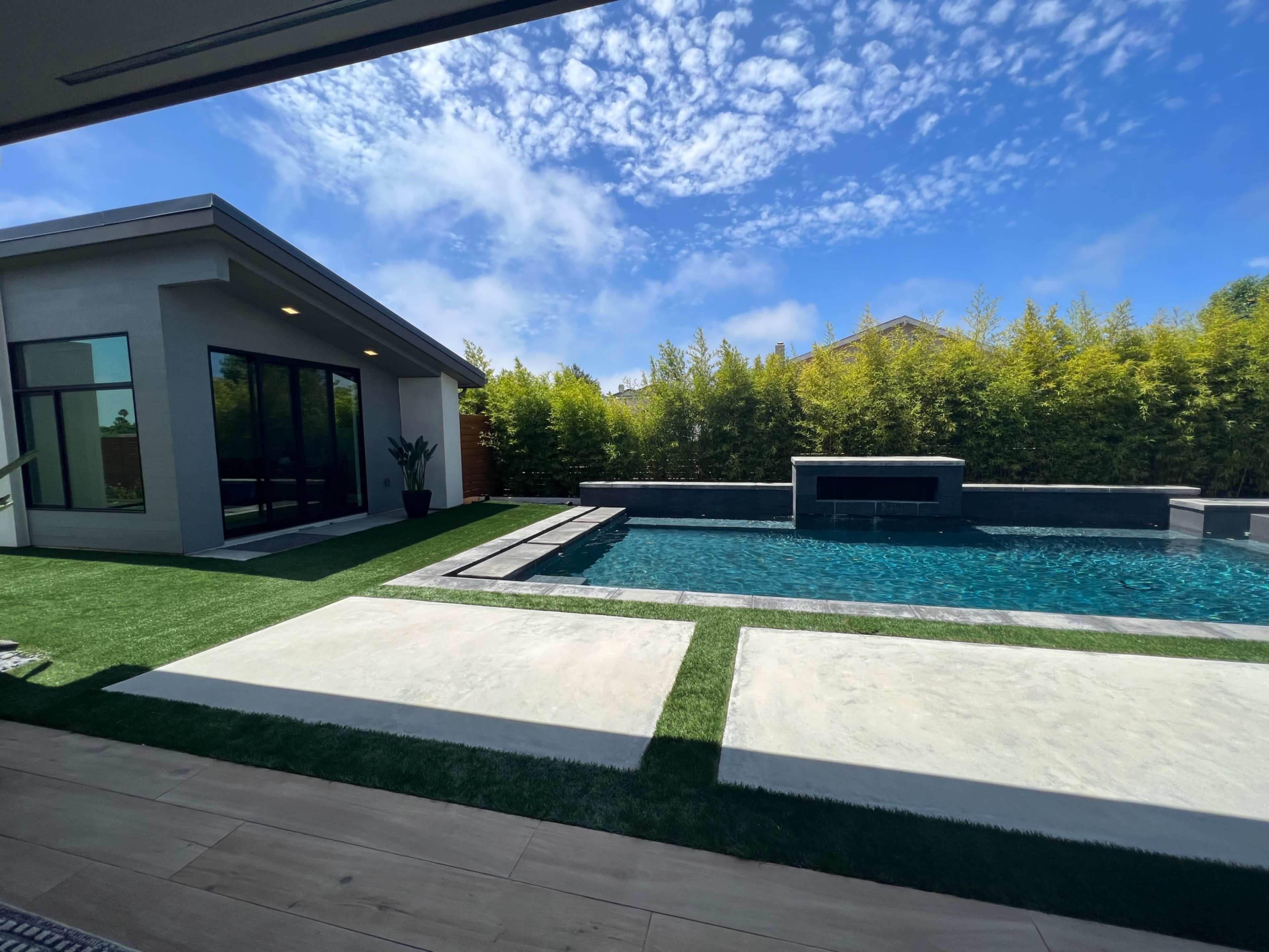 A modern backyard with a pool, concrete patio, and lush greenery surrounding a contemporary house under a blue sky.