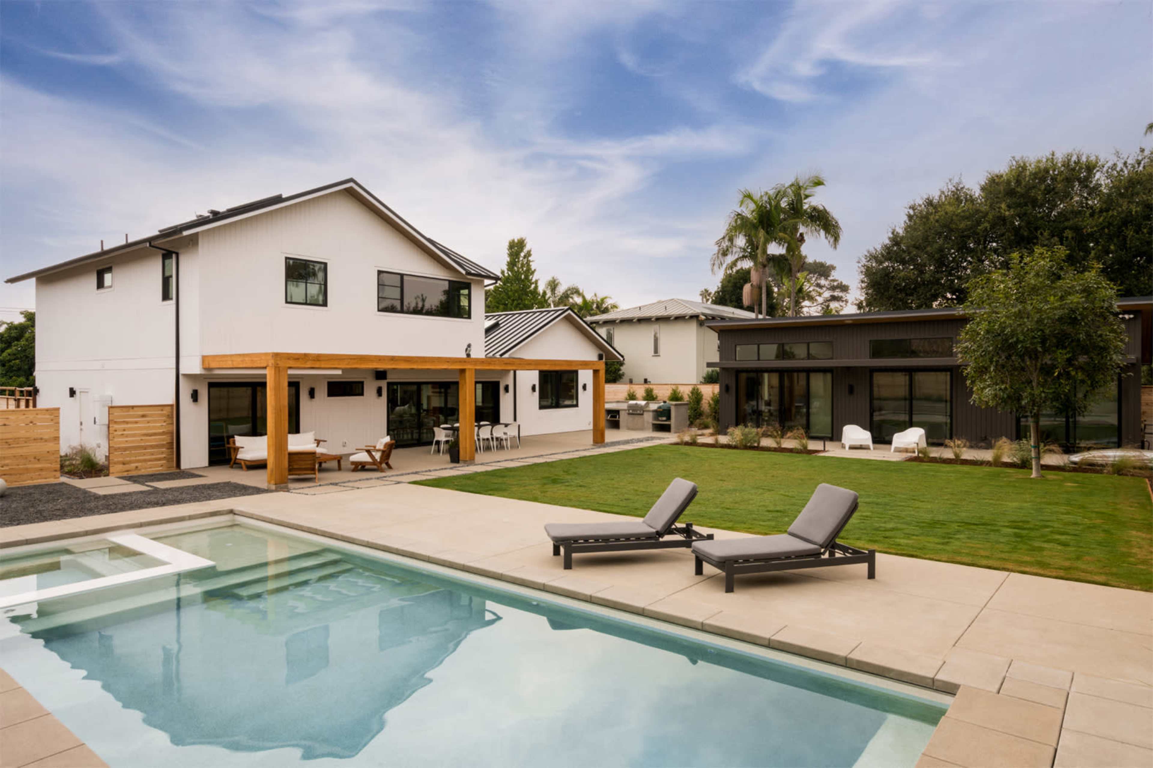 A modern backyard features a swimming pool, lounge chairs, and a stylish home with a wooden pergola and landscaped grass areas.