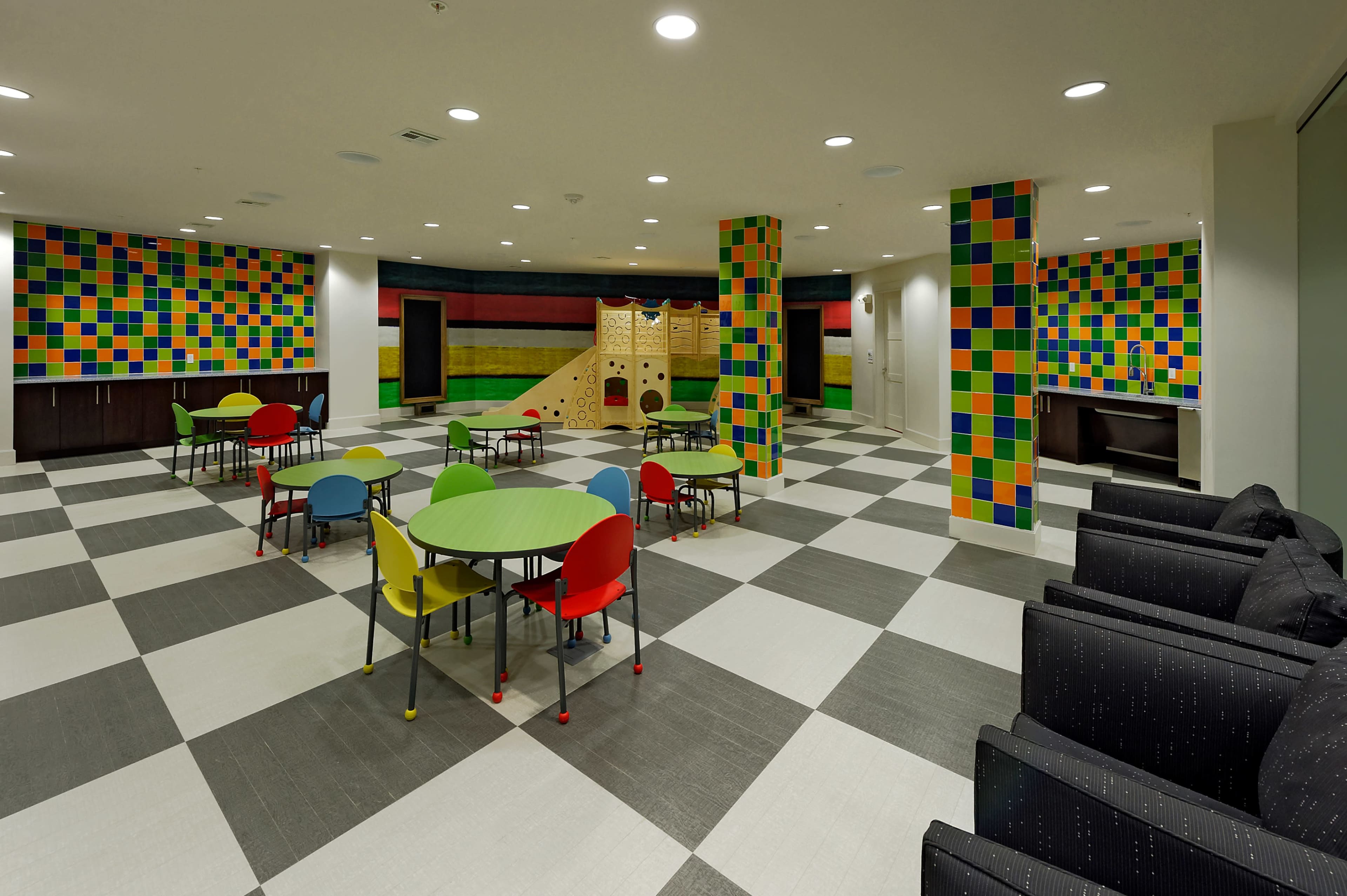 A brightly colored playroom with multicolored wall panels, green and red tables, and a climbing structure.