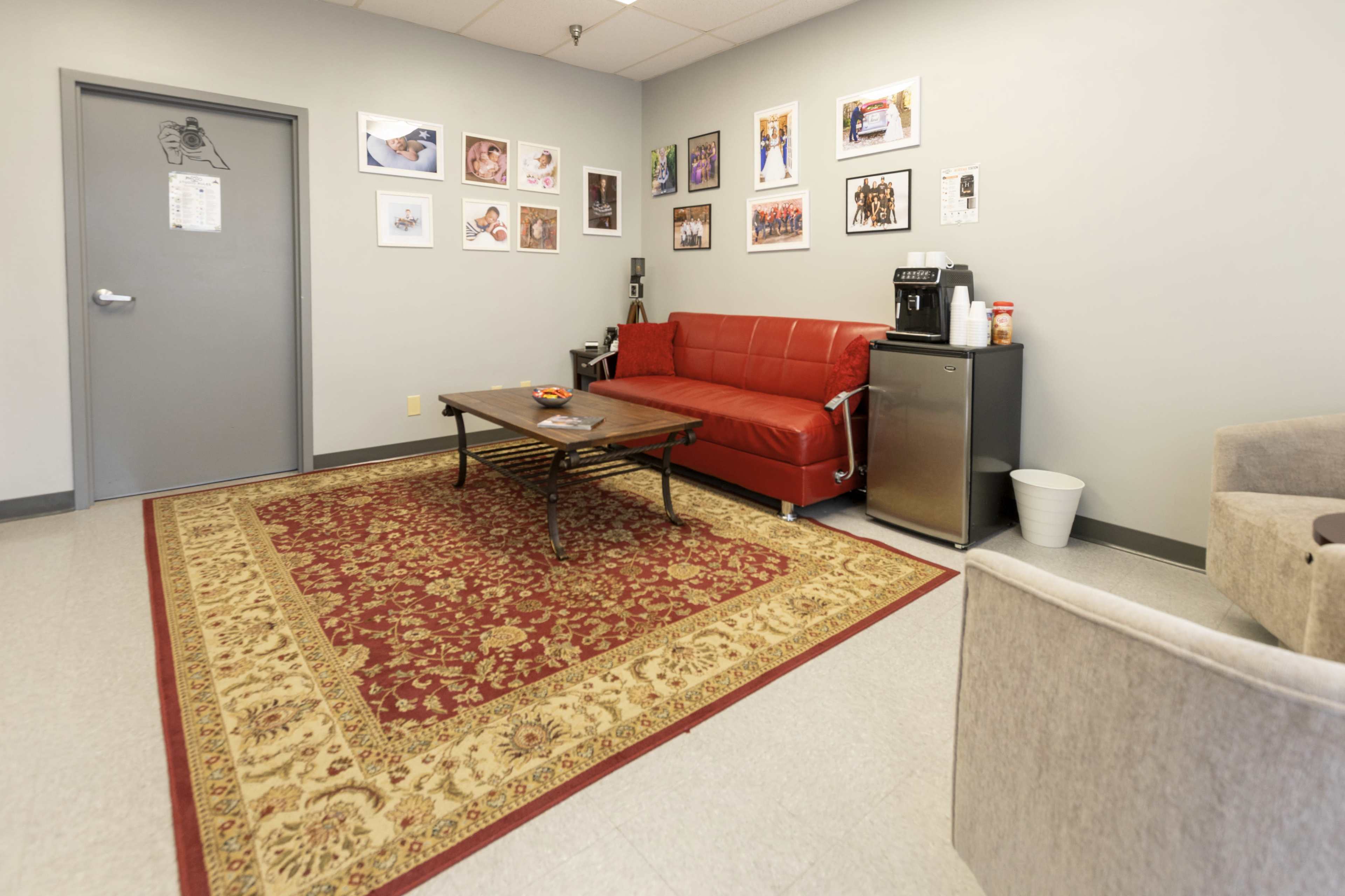 The image shows a waiting room with a red couch, a coffee station, photographs on the wall, and a patterned rug on the floor.