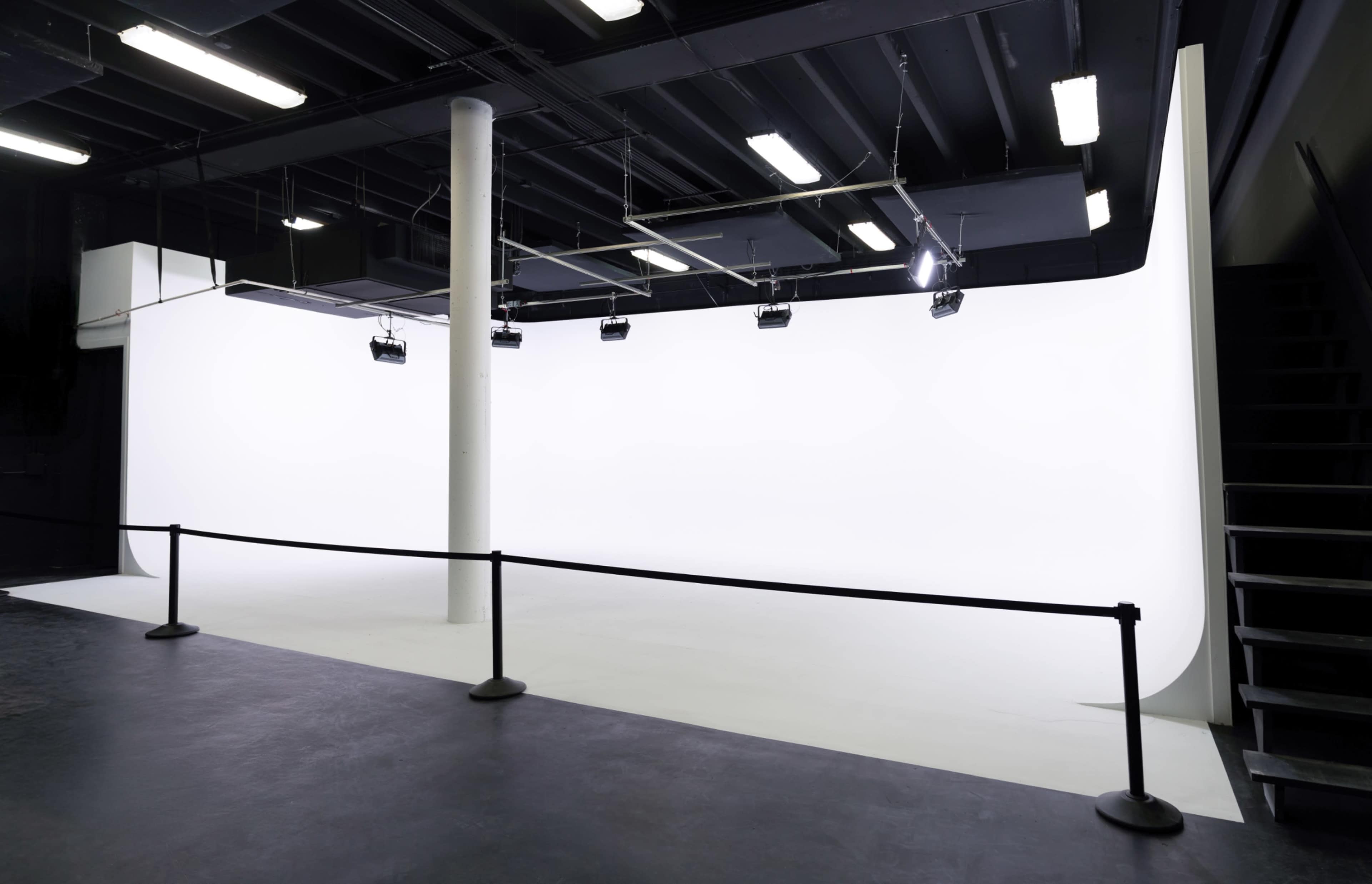 The image shows a spacious photography studio with a seamless white backdrop, lighting fixtures above, and a black barrier in front.