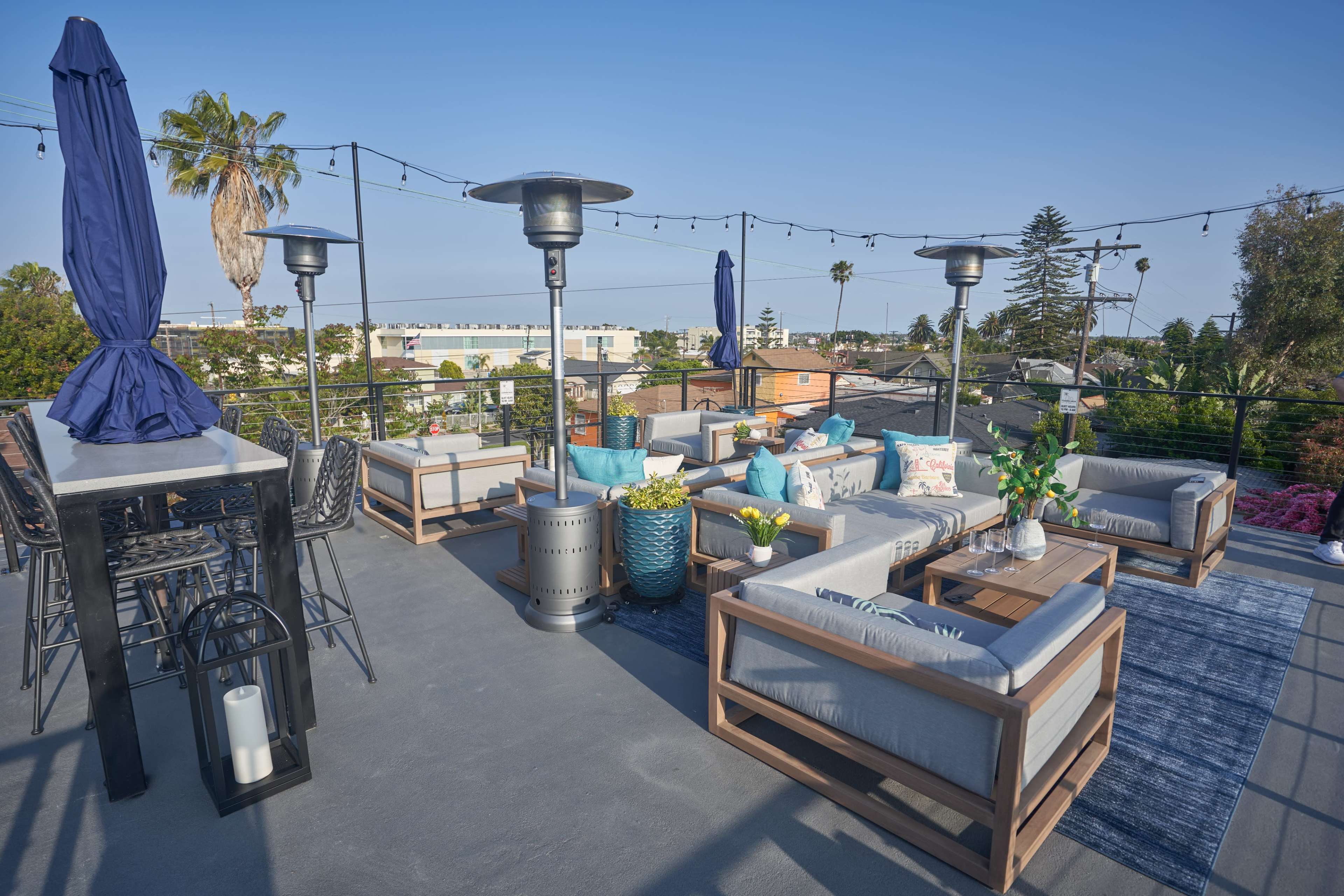 A rooftop terrace features outdoor seating with cushions, patio umbrellas, and potted plants against a clear sky.