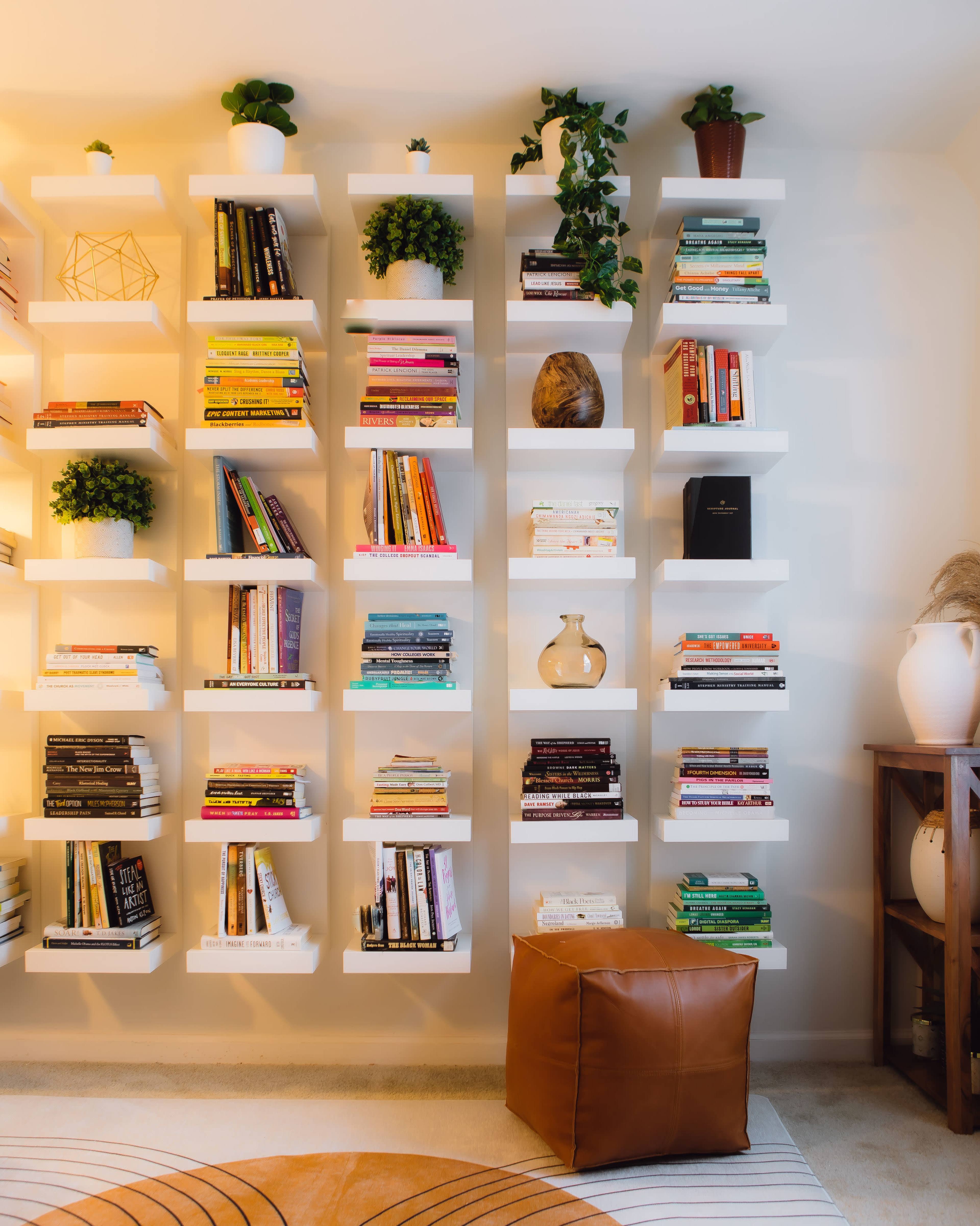 A white wall-mounted bookshelf displays a variety of books, plants, and decorative items, with a brown pouf and a rug on the floor.