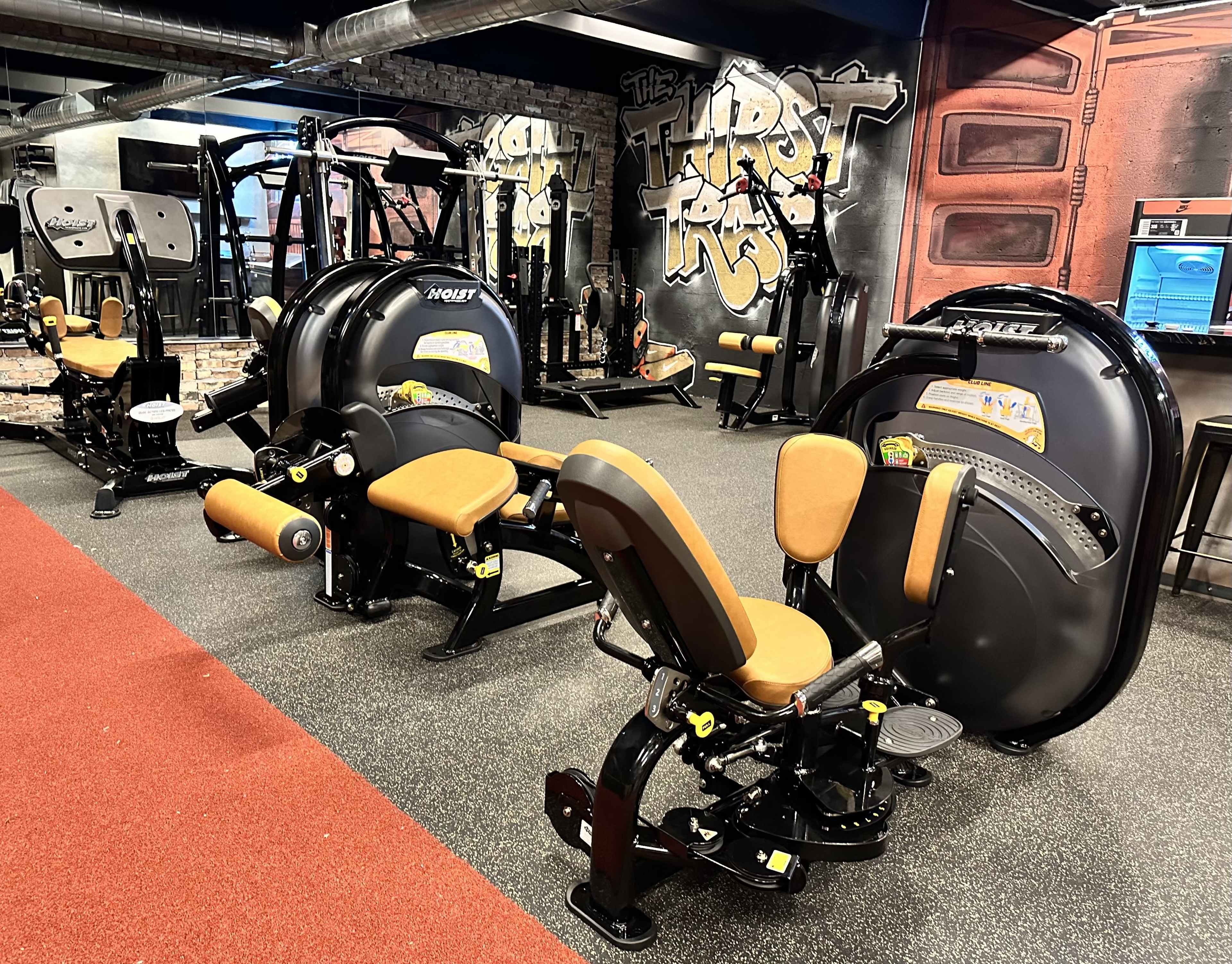 A gym interior featuring multiple exercise machines with padded seats arranged on a rubber floor.