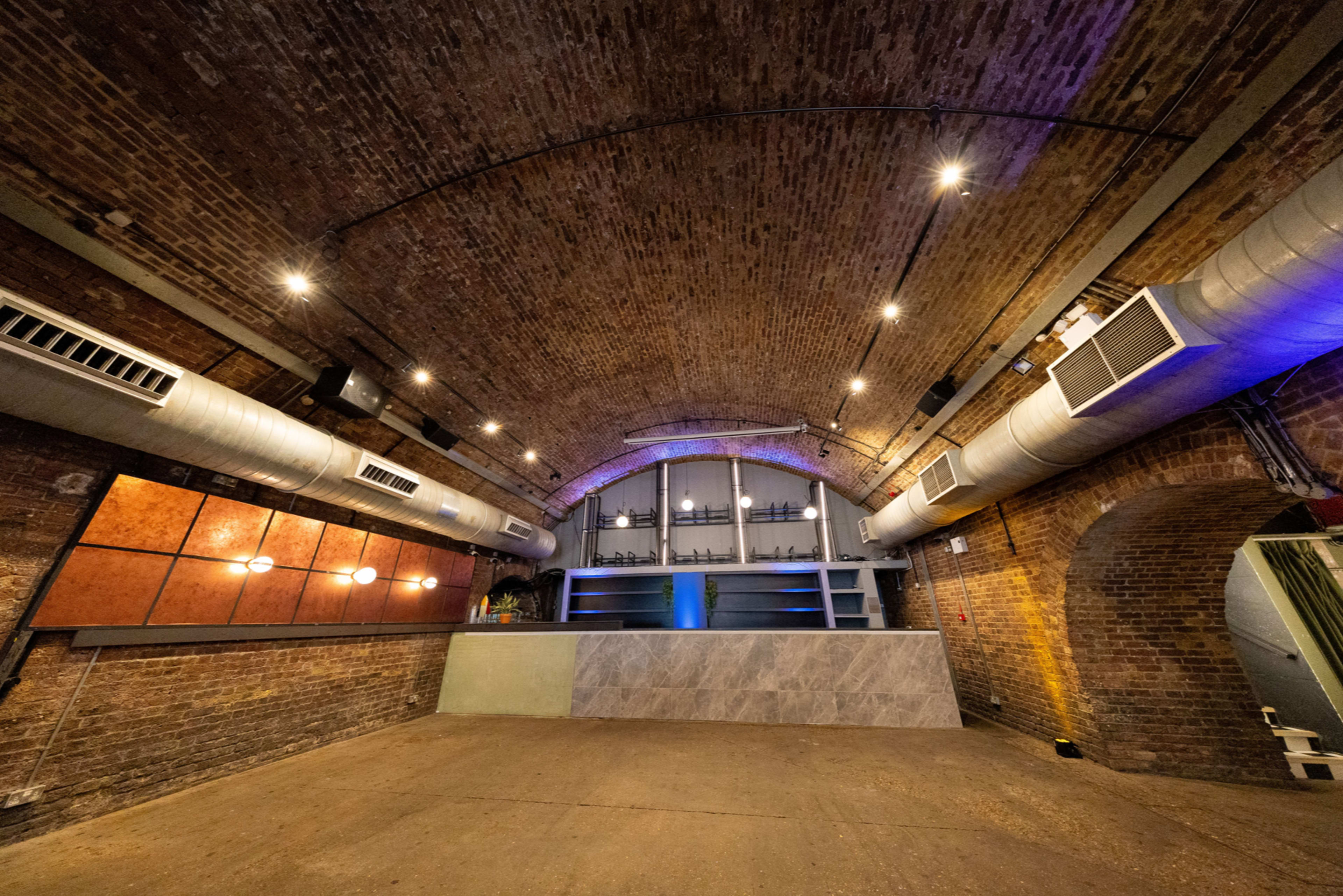 The image shows a spacious interior of a venue with a brick arch ceiling, equipped with a bar area featuring a sleek countertop and industrial-style lighting.