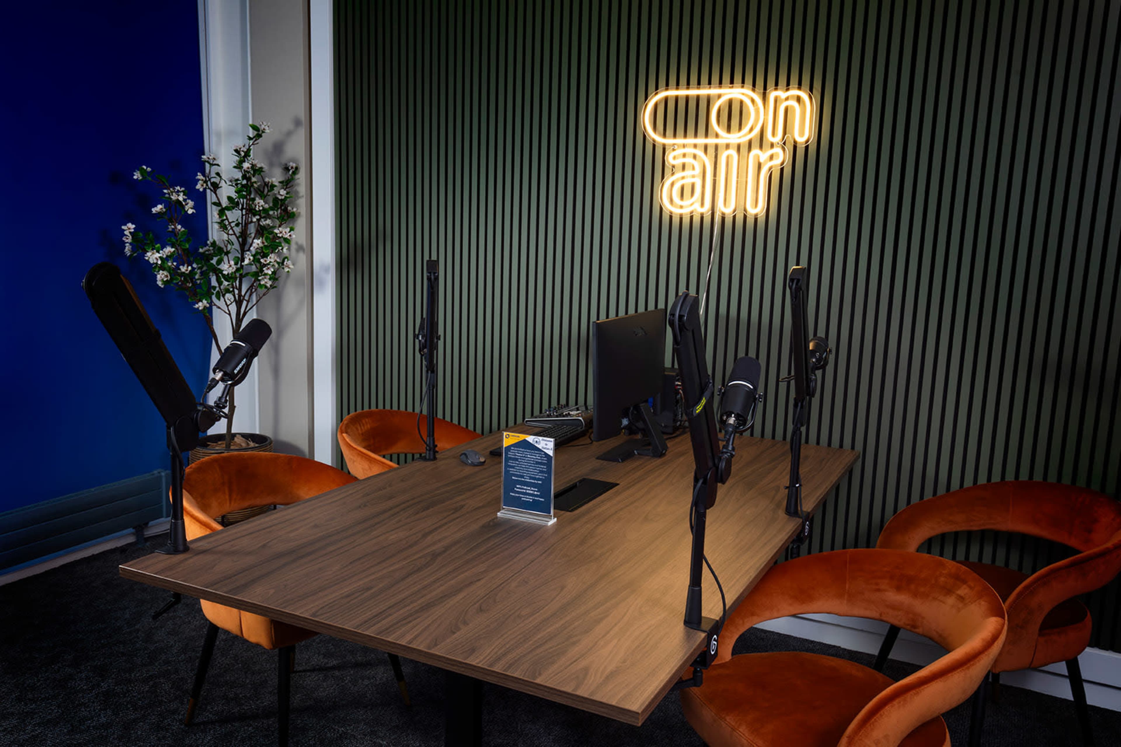 A modern podcast recording studio features a wooden table with four chairs, a computer, and multiple microphones arranged around it, illuminated by a neon "on air" sign.
