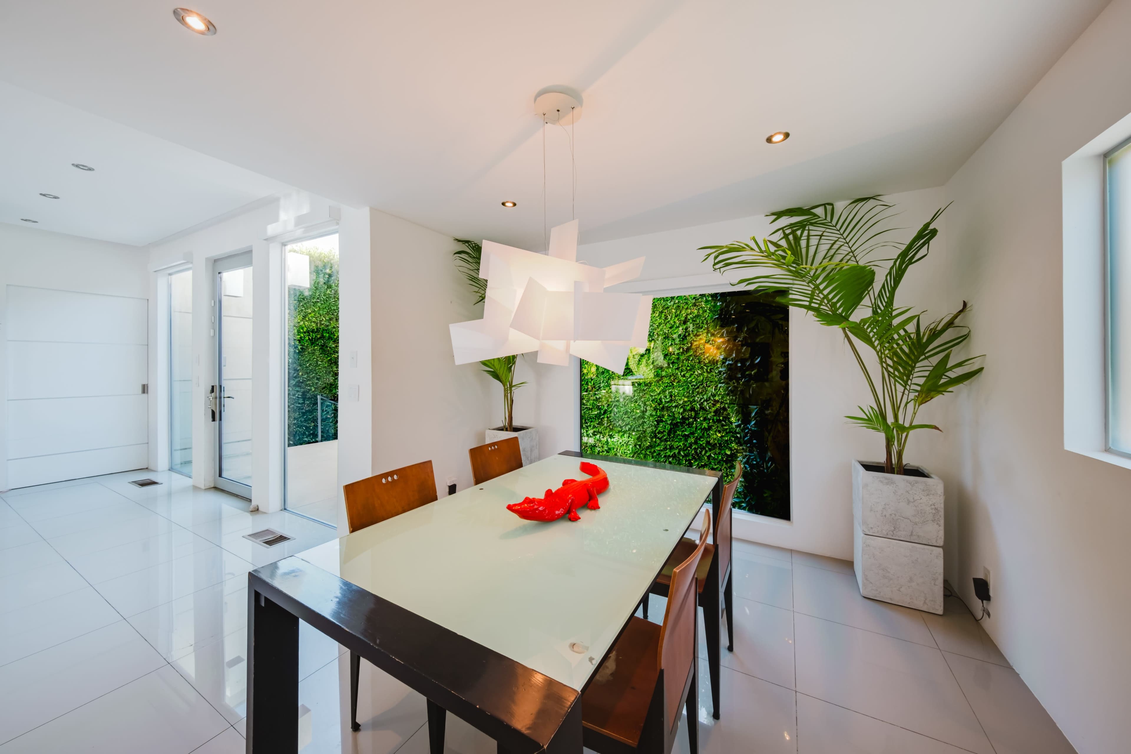 A modern dining area features a glass table surrounded by wooden chairs and is illuminated by a unique pendant light, with large windows revealing greenery outside.