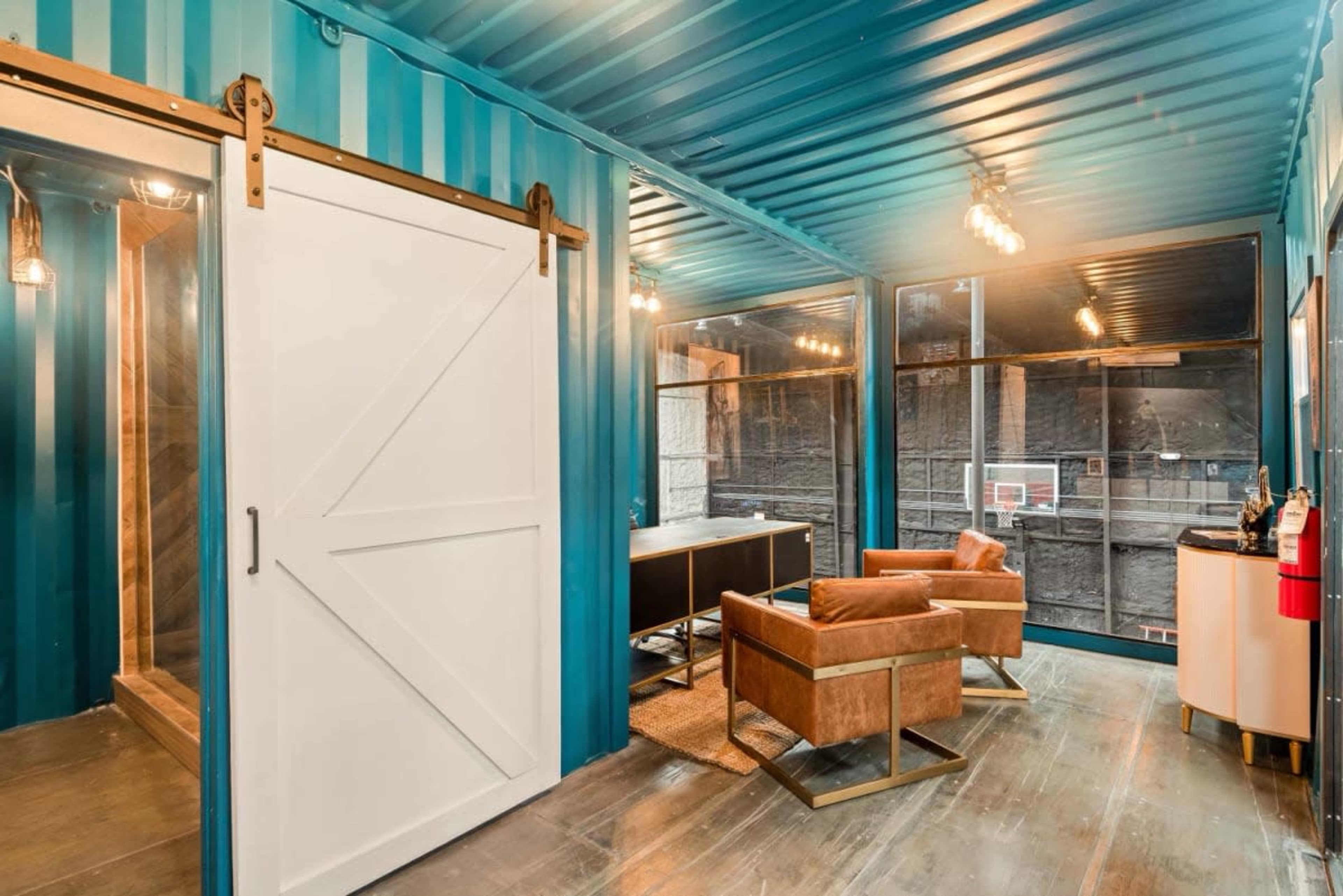 The image shows a modern office space with teal walls, a sliding barn door, large windows, and two brown leather chairs.
