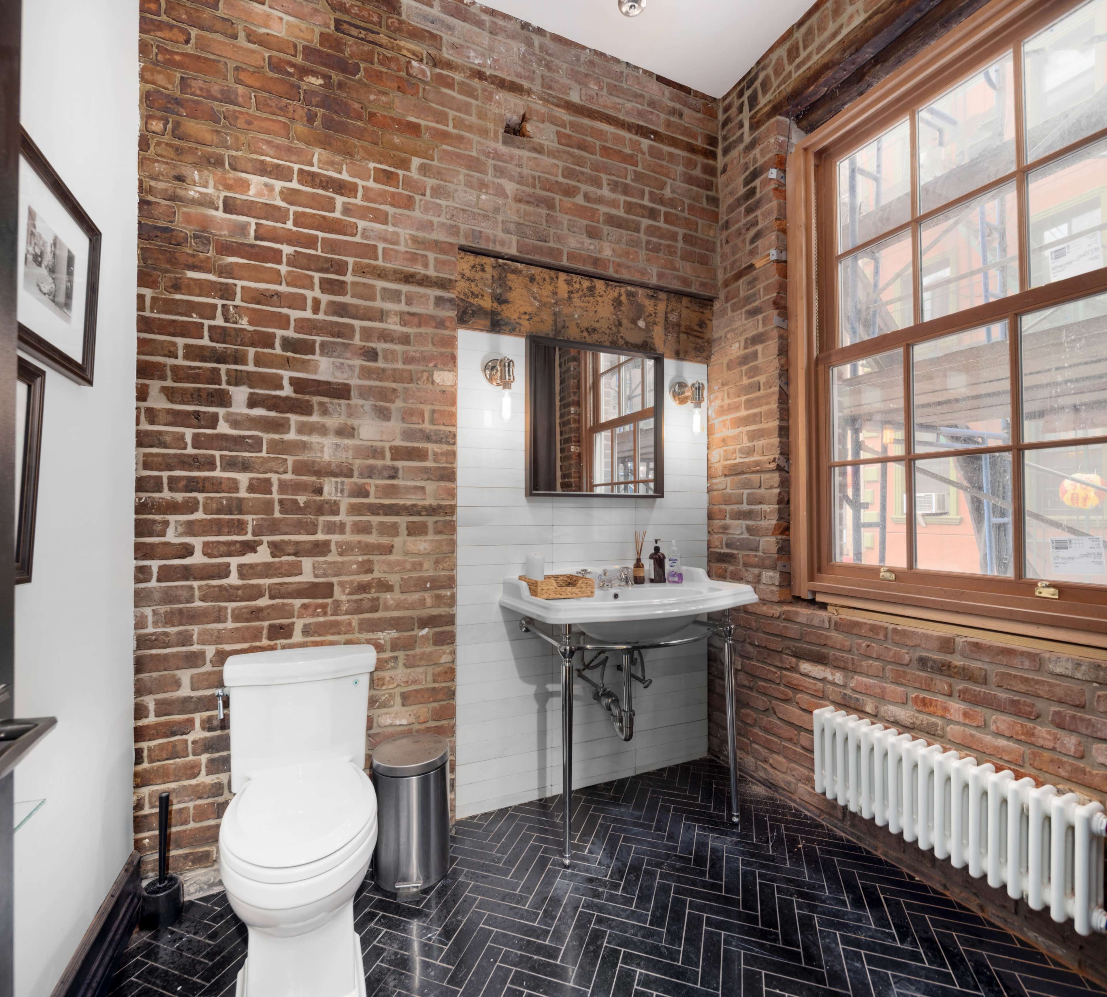A bathroom features exposed brick walls, a wall-mounted sink with a mirror, and a toilet, alongside a white radiator and dark herringbone flooring.