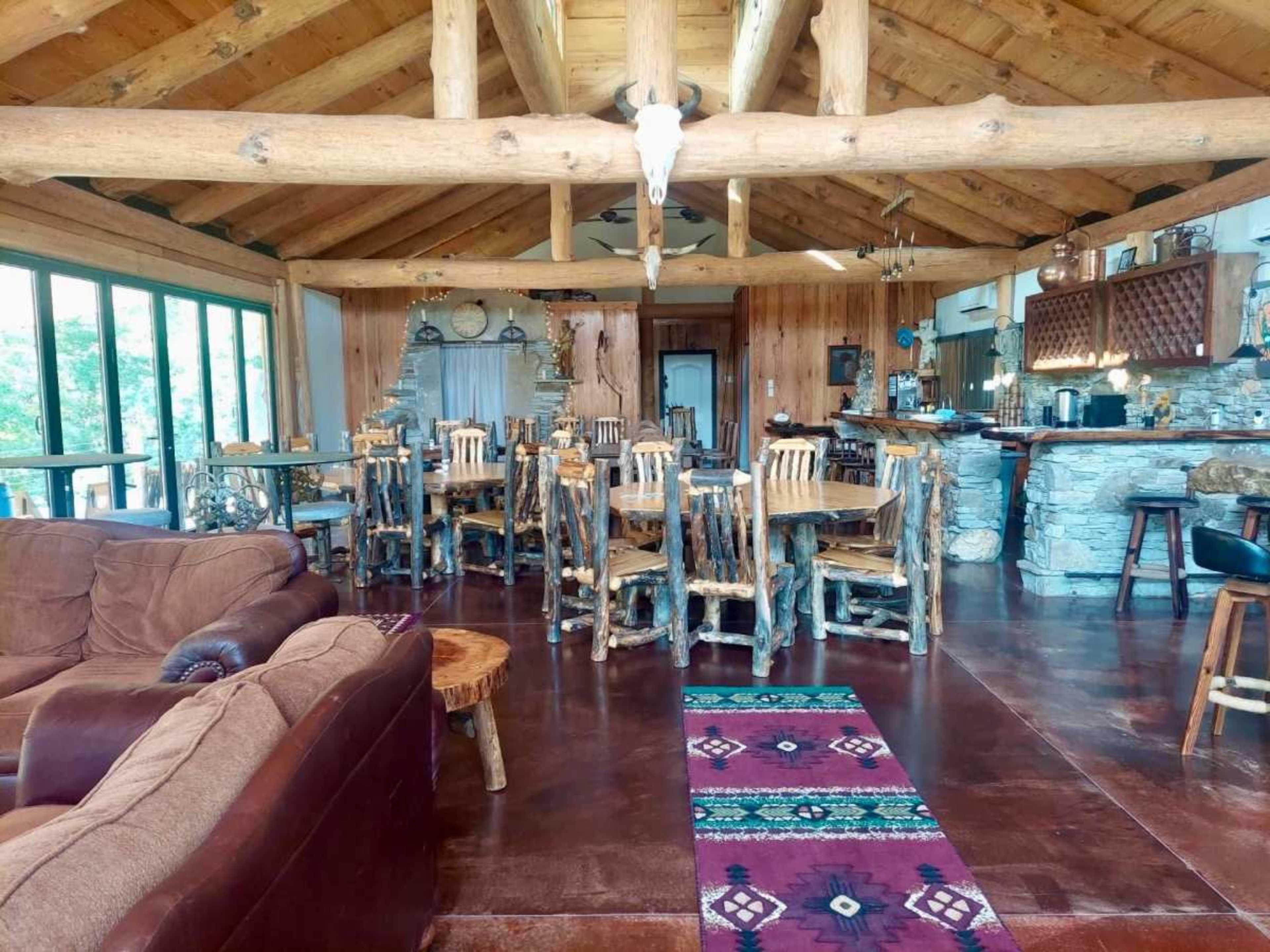 The image shows a rustic interior of a spacious lodge with wooden beams, a dining area featuring a large table surrounded by log-style chairs, and a cozy seating area.