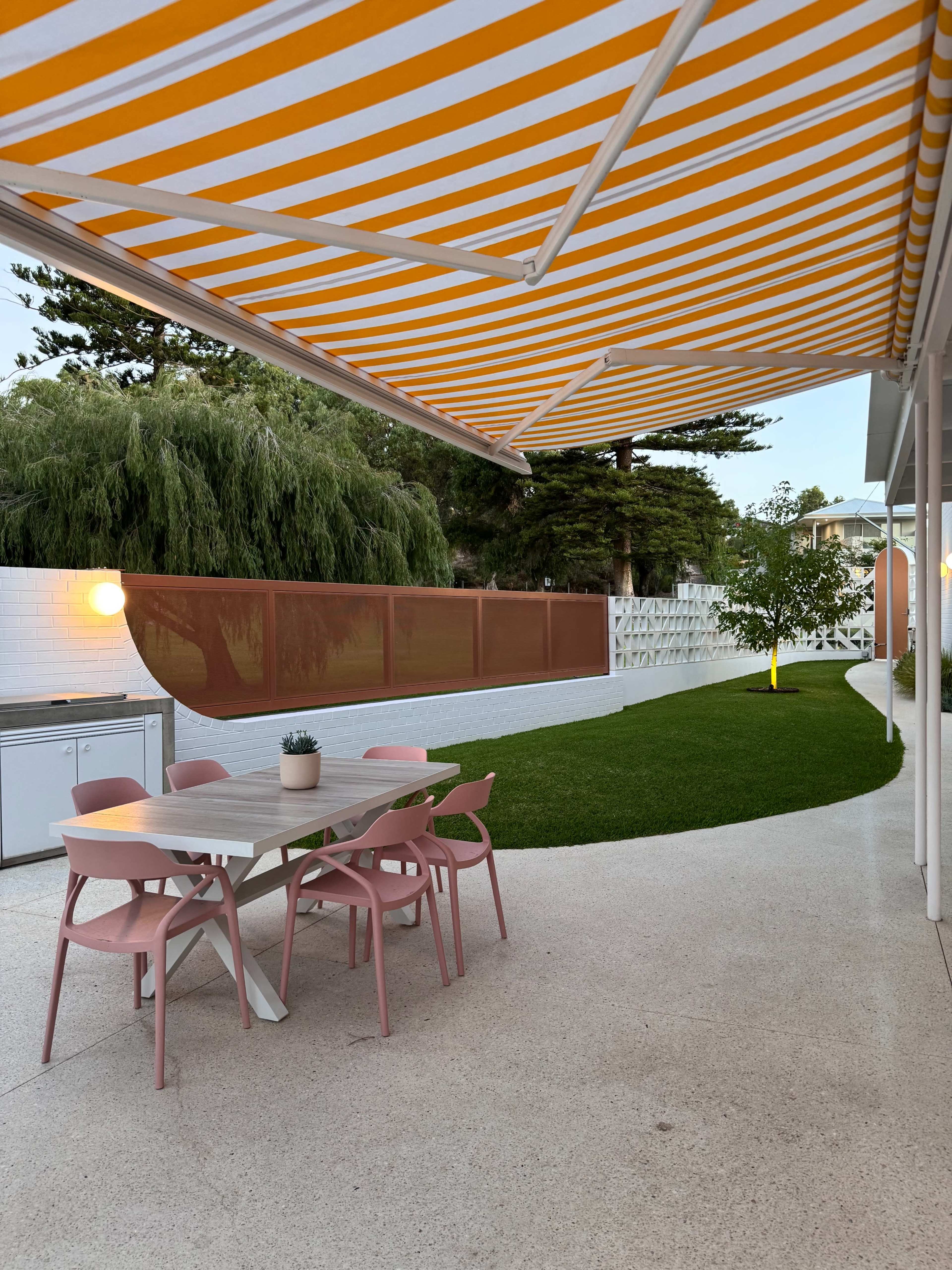 A patio area features a table with pink chairs, an outdoor kitchen, and a neatly maintained lawn beneath a striped awning.