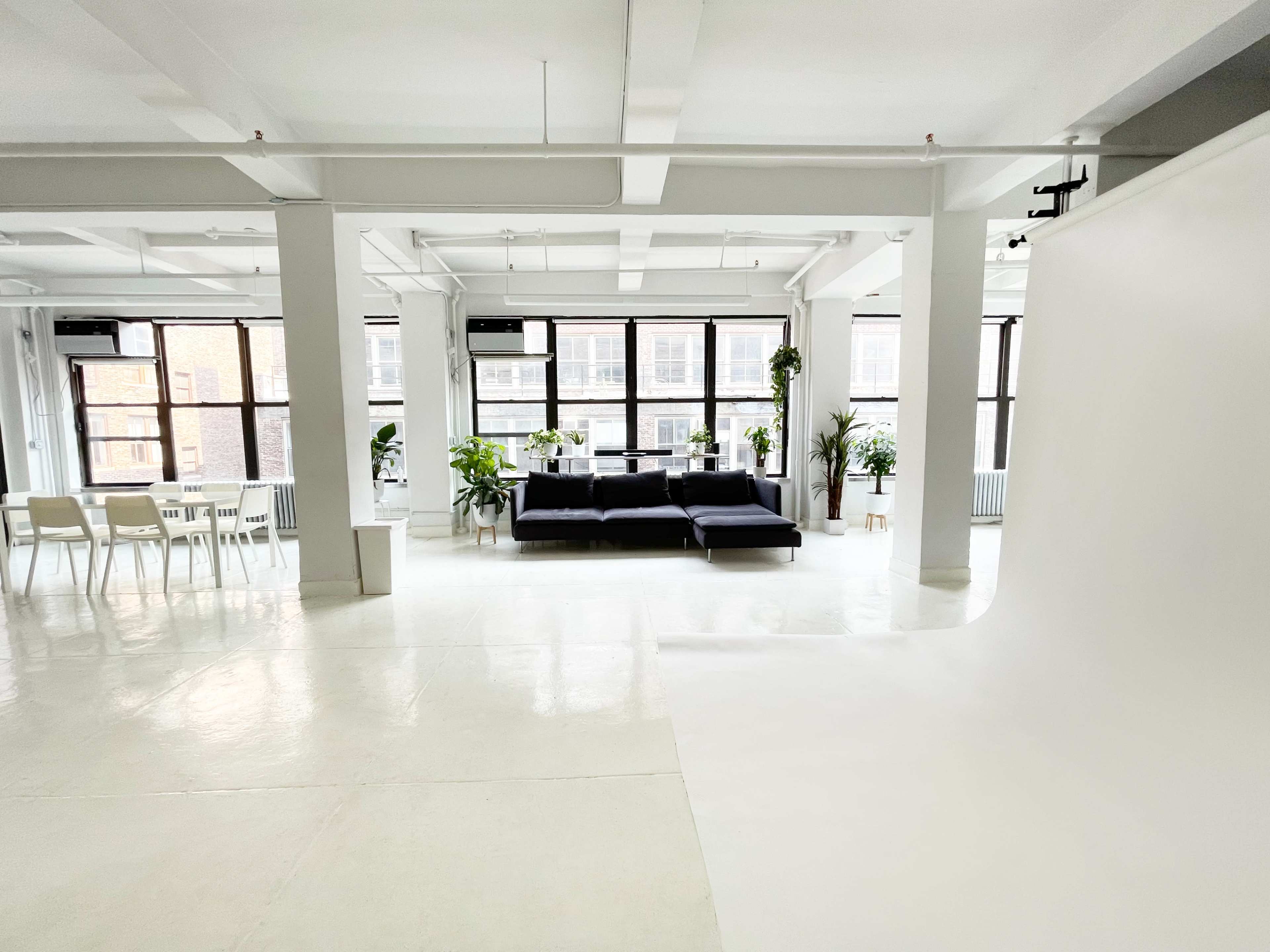 A bright, open space with large windows, a black sofa, and a dining area with white chairs and a table, alongside potted plants.