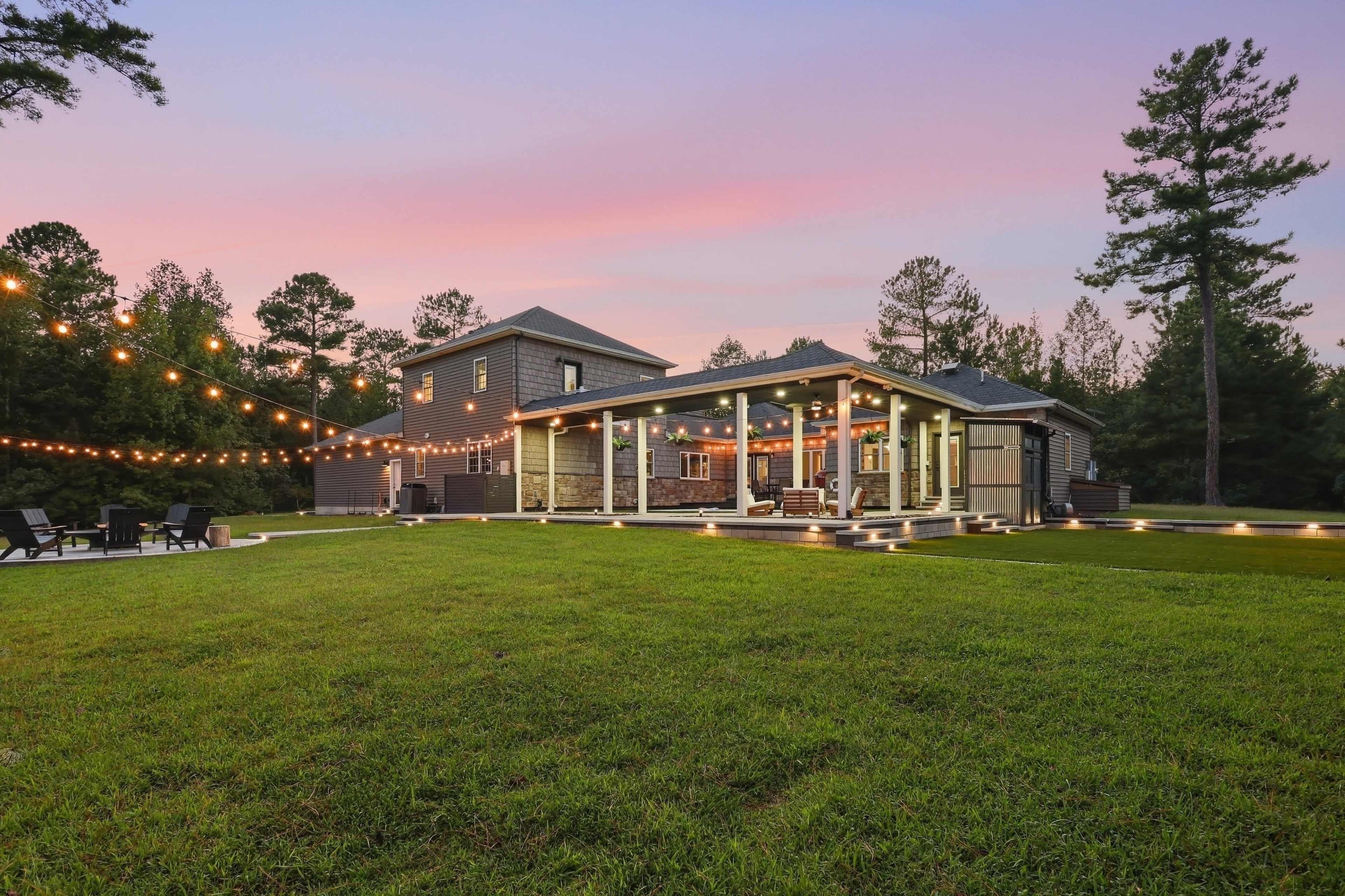 A modern house with a patio, string lights, and a well-manicured lawn is set against a colorful sunset sky.