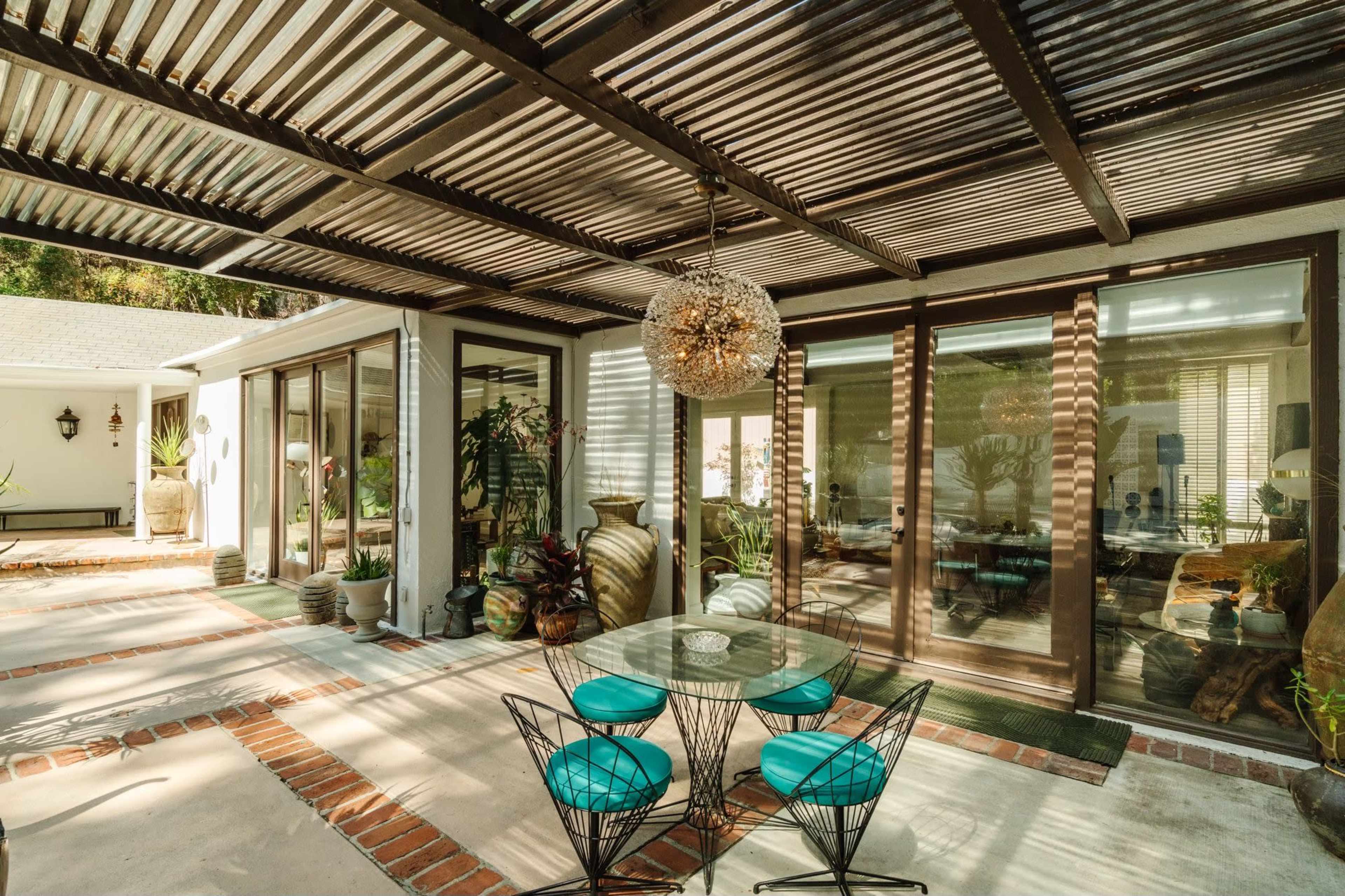 The image shows a covered outdoor patio area featuring a round glass table with turquoise chairs, surrounded by potted plants and decorative items.