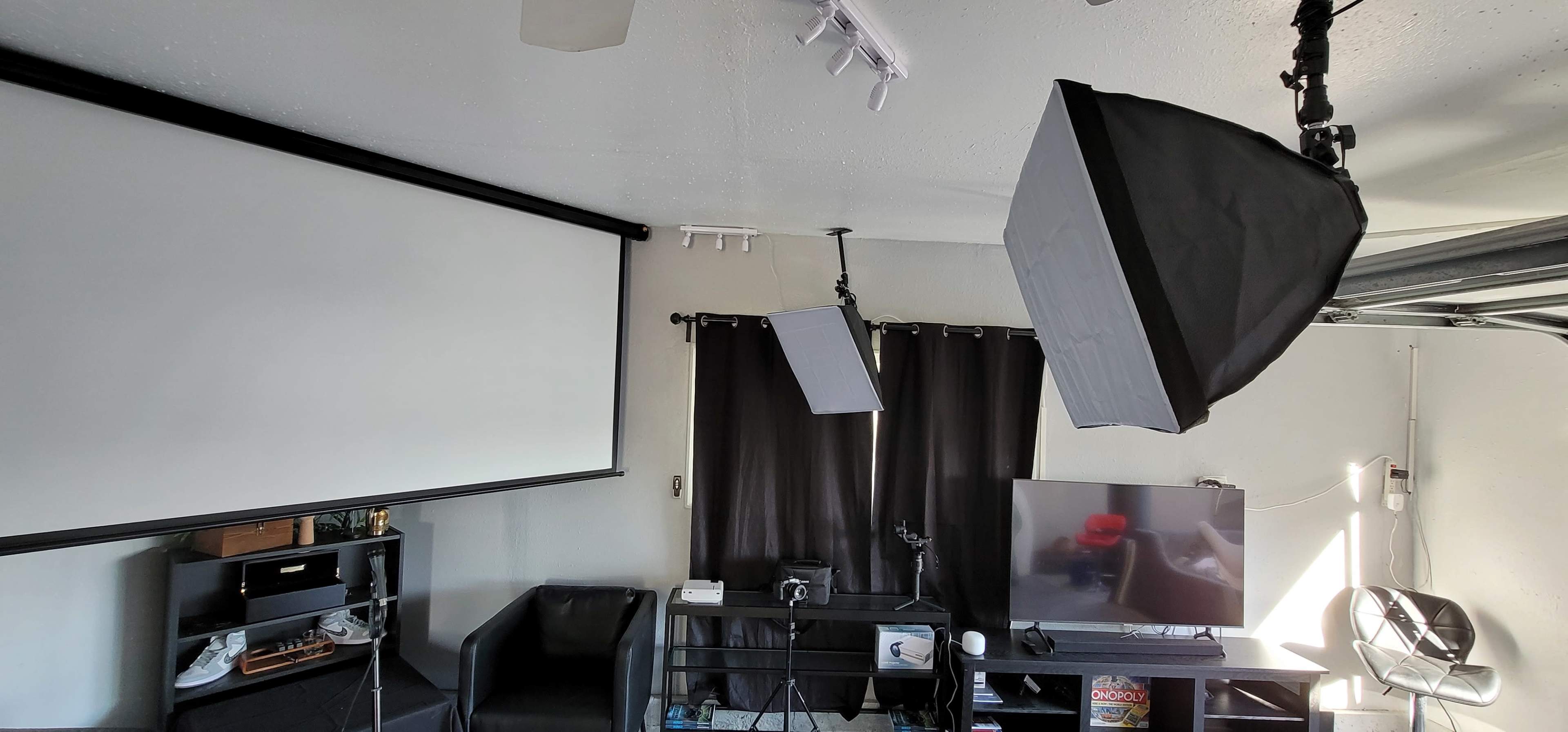 The image shows a modern living room setup featuring a large flat-screen TV, black curtains, a projector screen, and softbox lighting equipment.