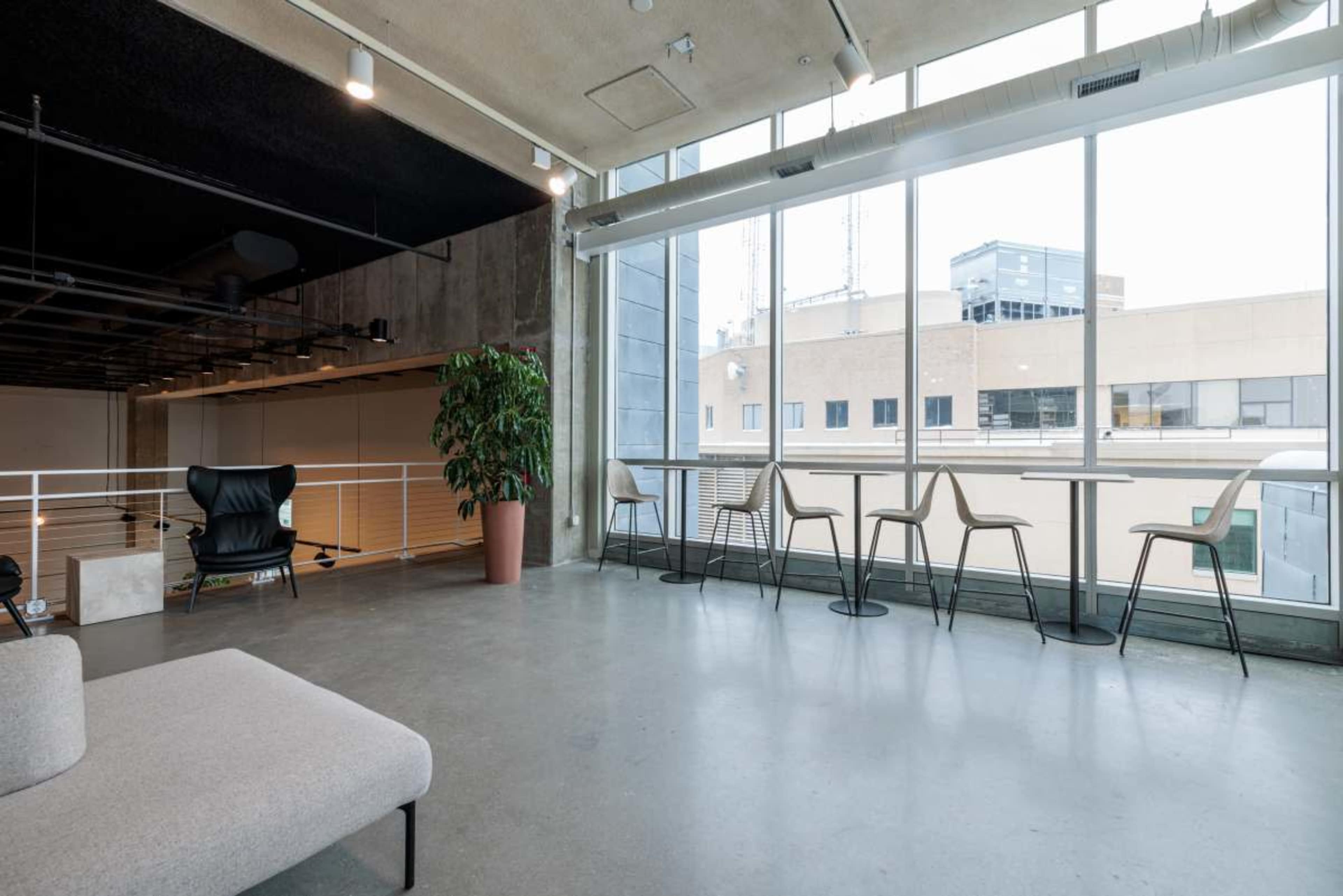 The image depicts a modern workspace with a concrete floor, large windows allowing natural light, and minimalistic furniture including a gray sofa and several chairs around small tables.