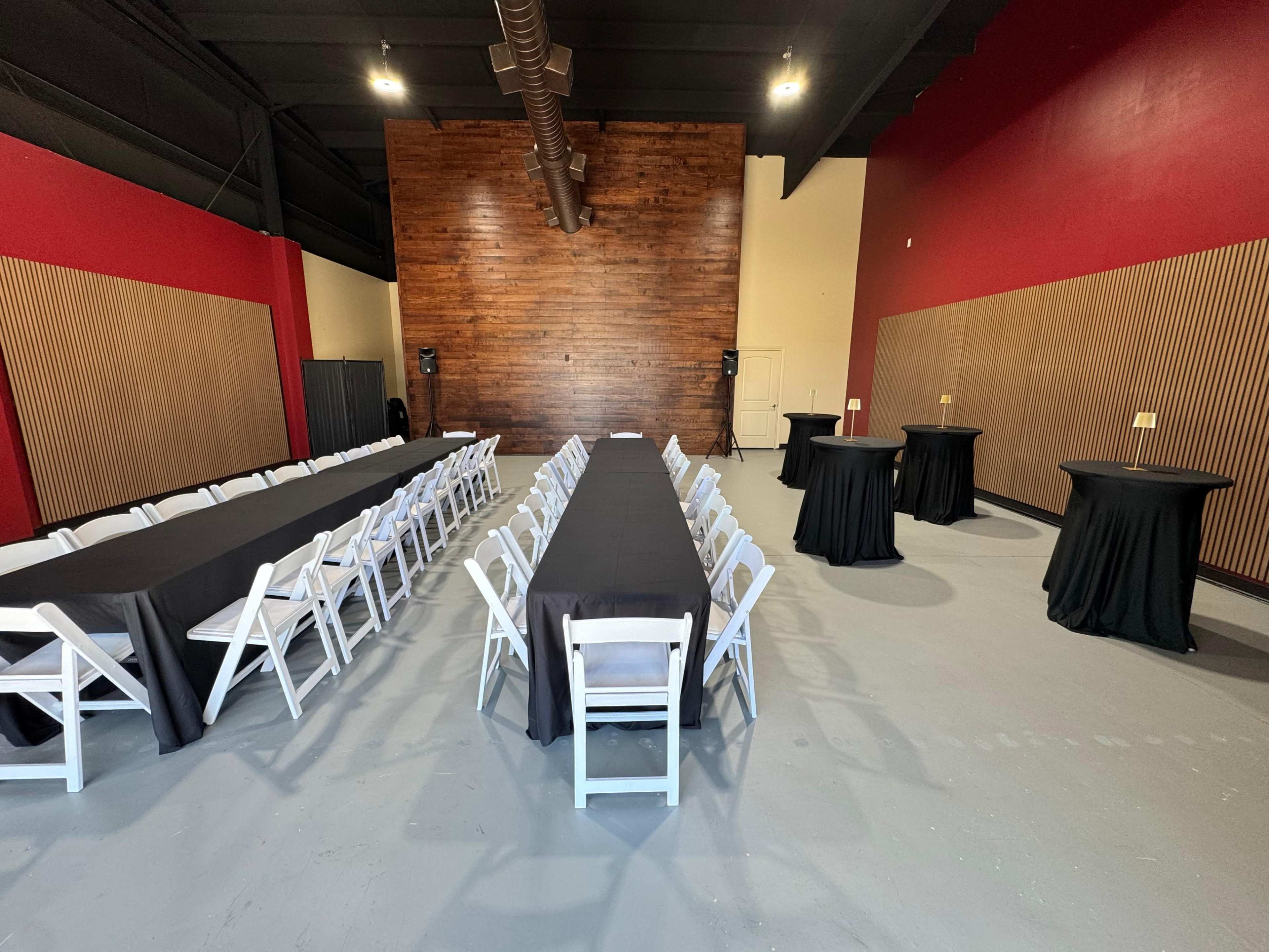 The image shows a spacious indoor venue set up for an event, featuring several tables covered with black cloth and arranged with white folding chairs along the sides.