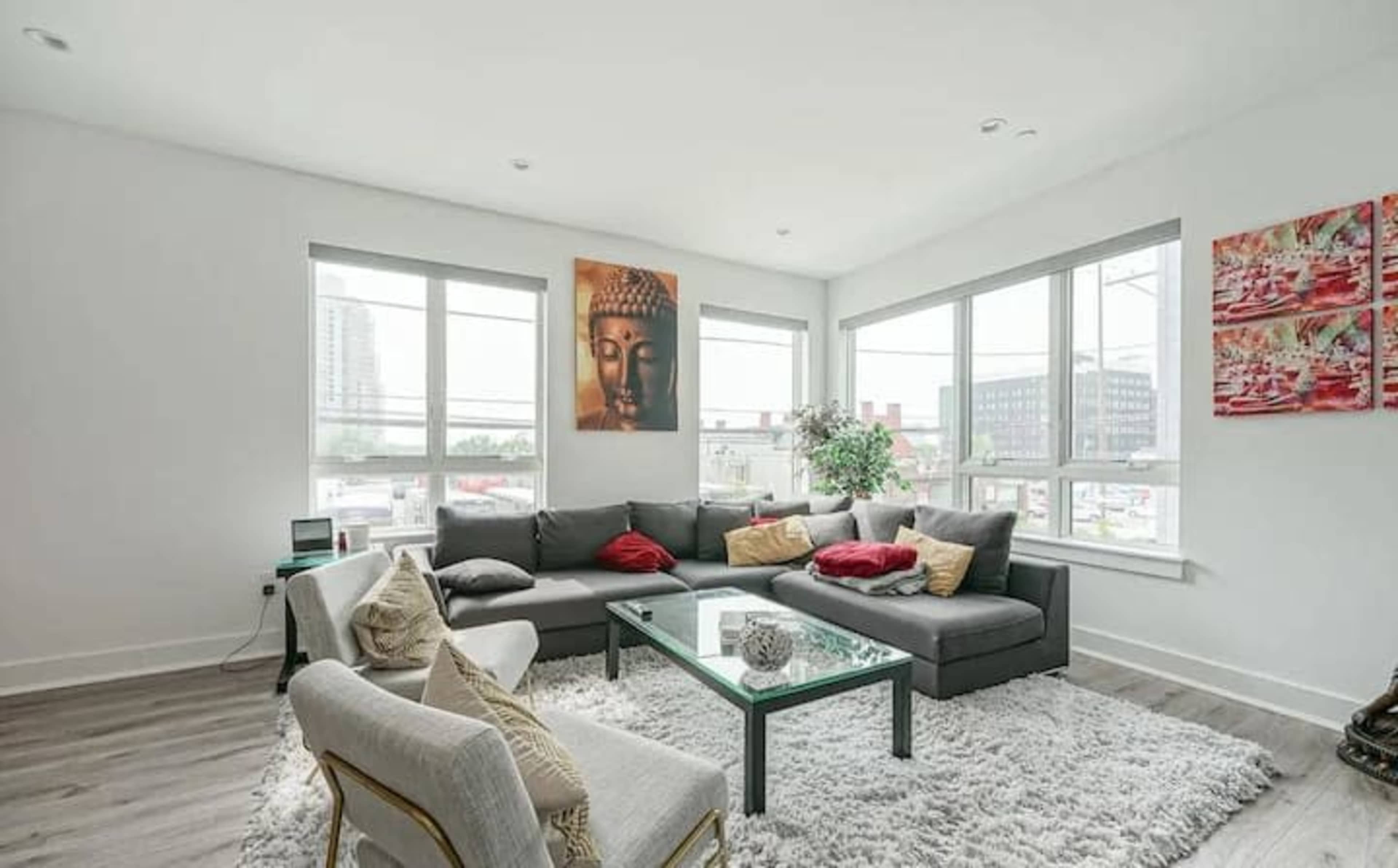 The living room features a sectional sofa with decorative pillows, a glass coffee table on a plush rug, and large windows that provide natural light.