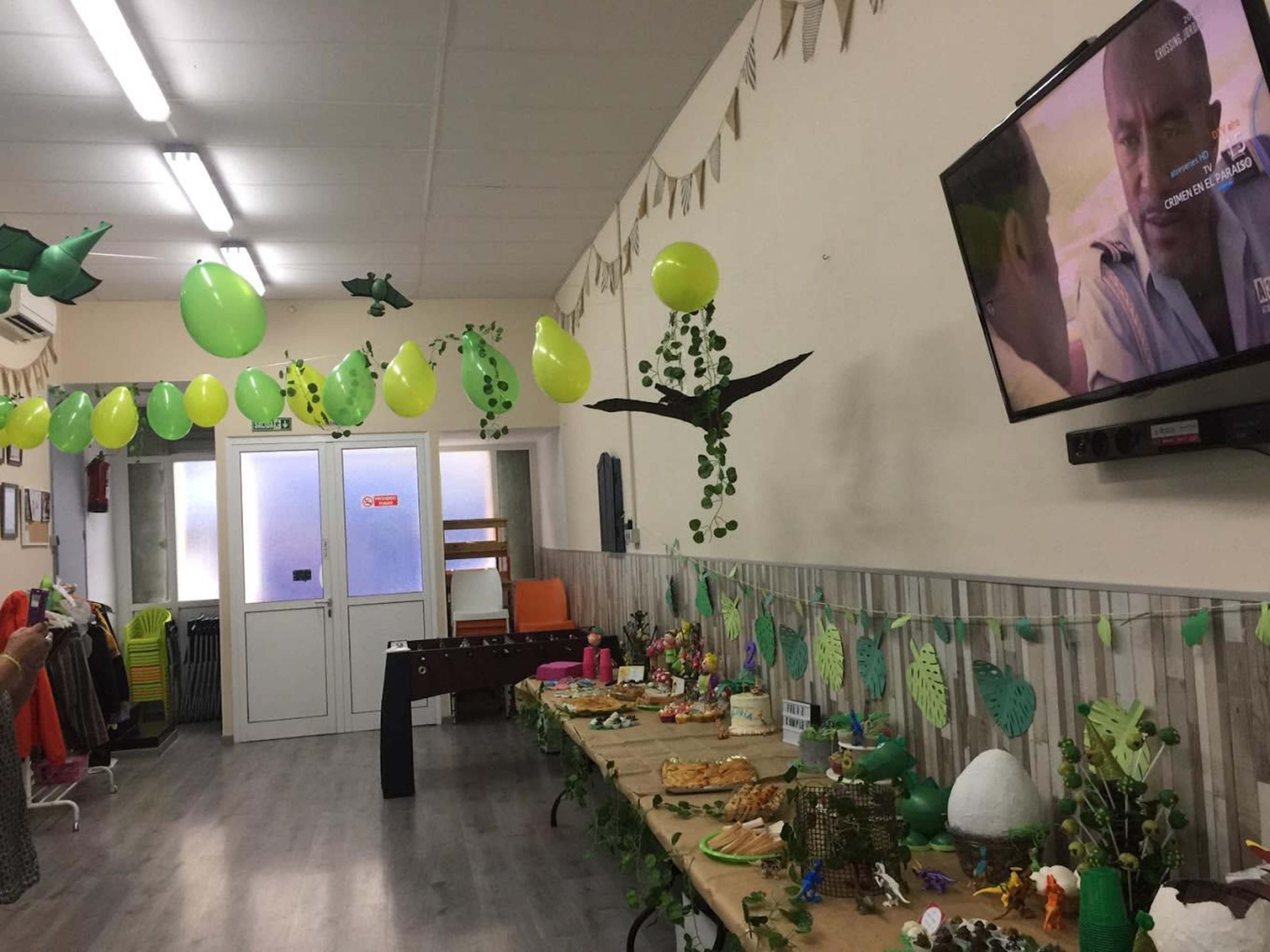 A decorated room features a table laden with food, green balloons, and plants hanging from the ceiling, while a television displays a scene from a show.