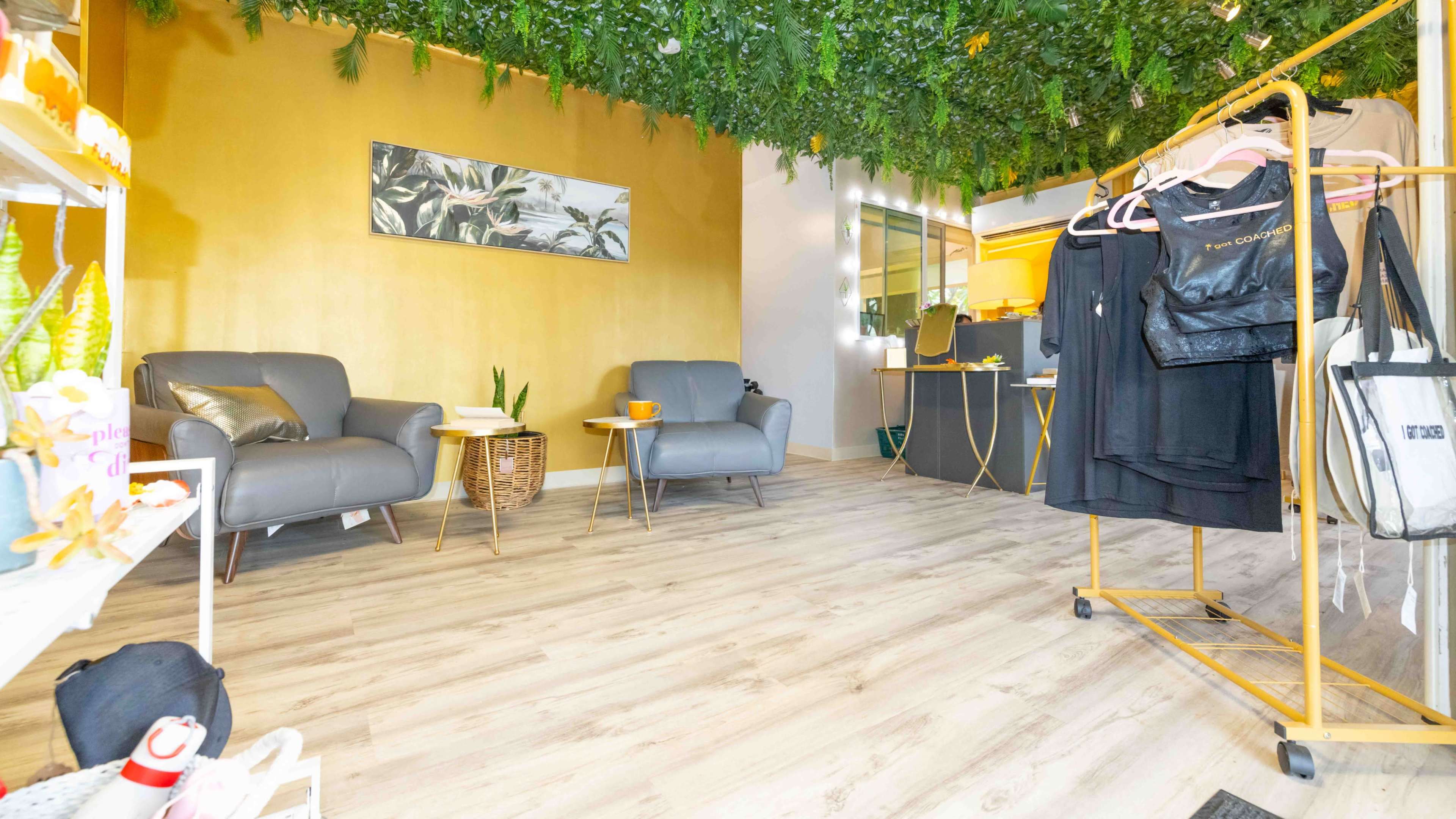 A cozy boutique interior features gray seating, a wall adorned with tropical art, and clothing racks amidst lush greenery.