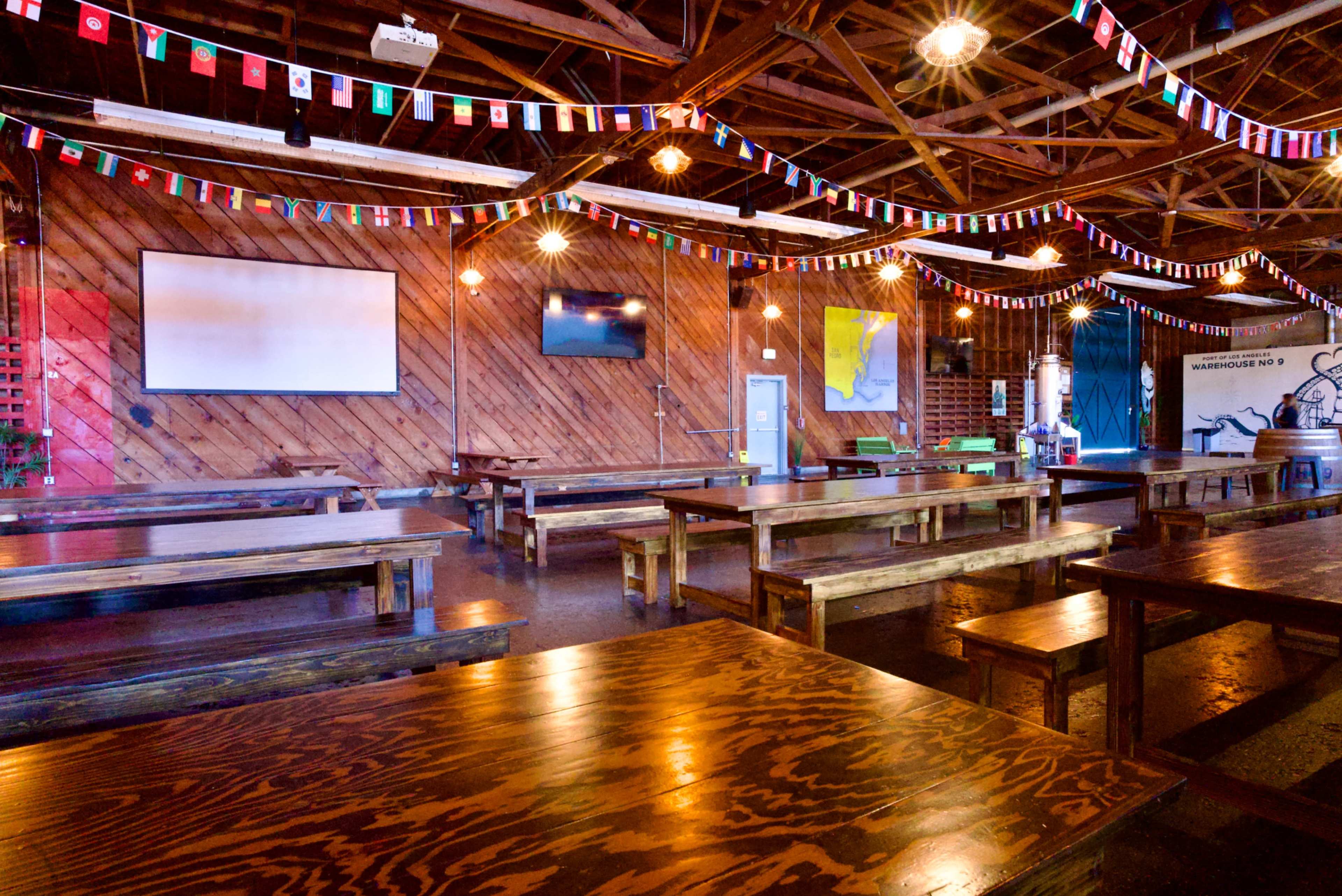 The image shows a spacious indoor venue with wooden benches and tables, adorned with colorful flags strung across the ceiling.