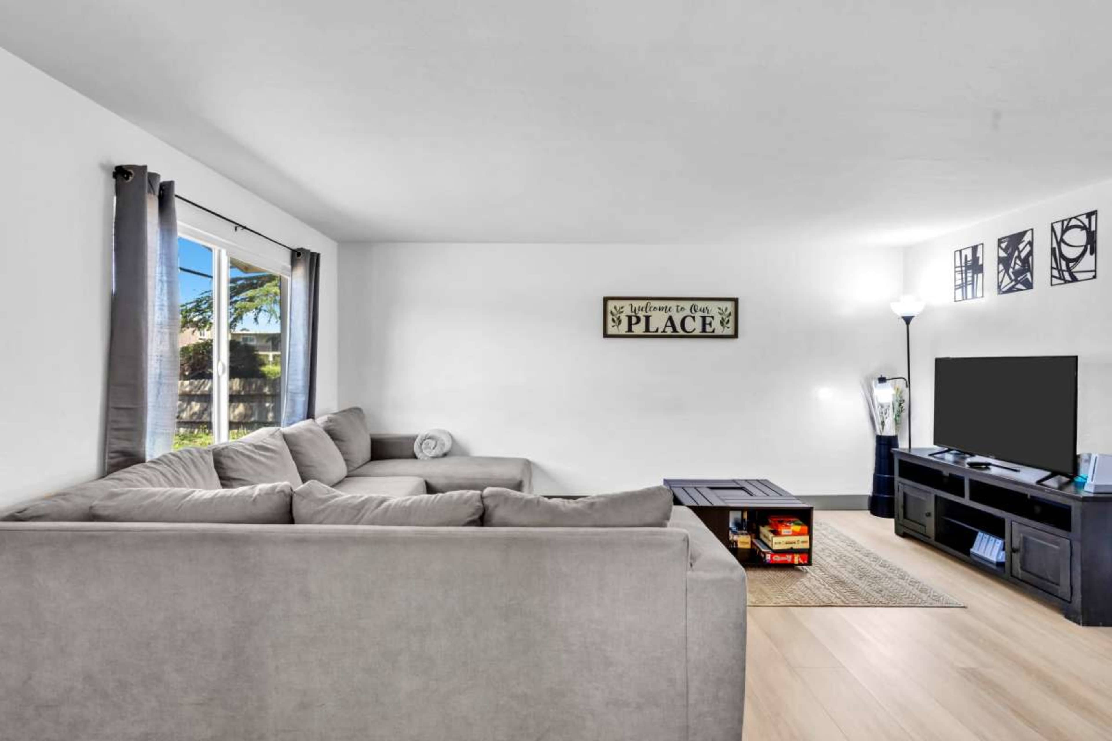 The image shows a living room with a light-colored sectional sofa, a television stand holding a TV, and a small coffee table, all set against a white wall with natural light coming through a window.