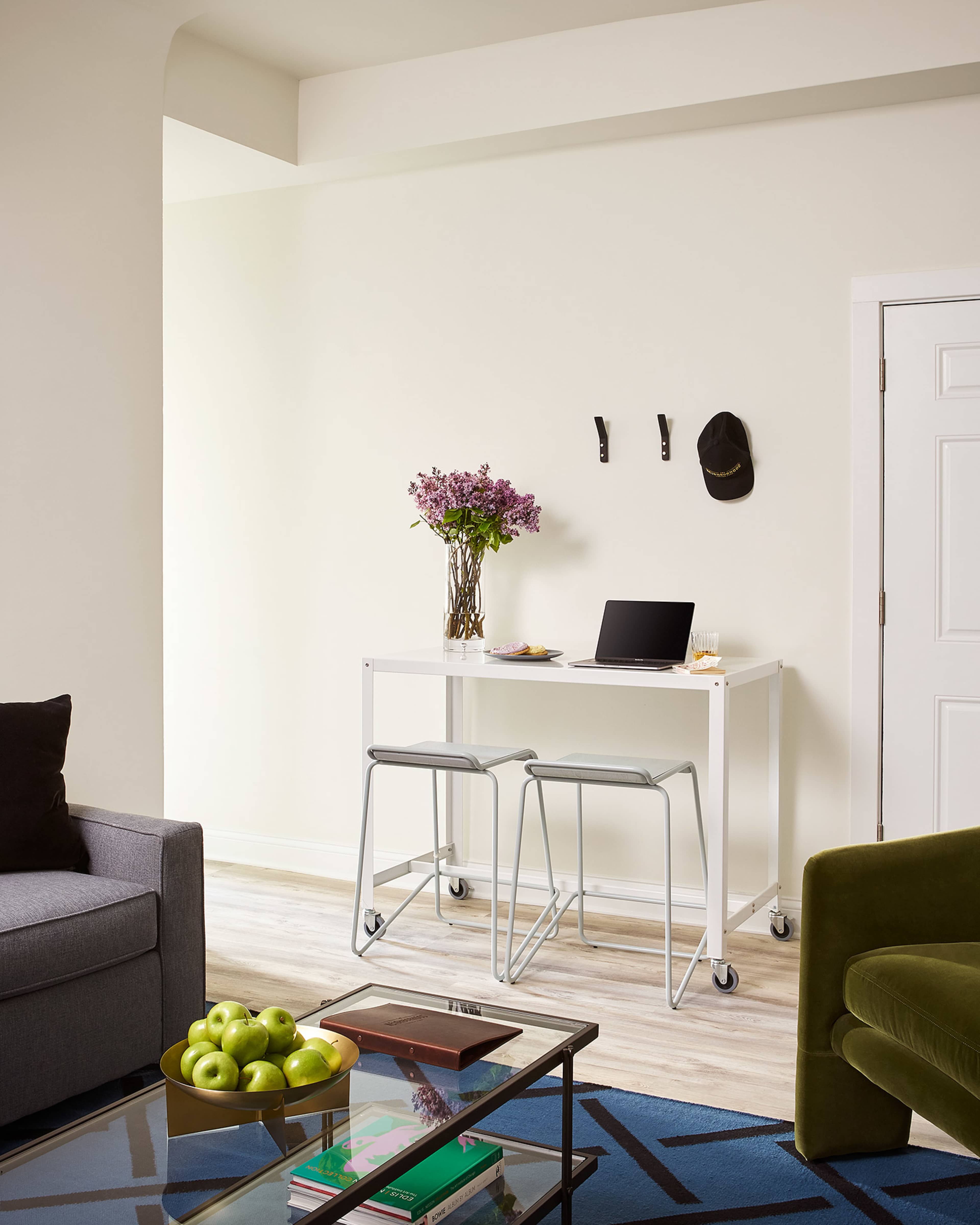 A small work area features a minimalist table with a laptop, a vase of flowers, and two stools, set against a light-colored wall in a cozy room.