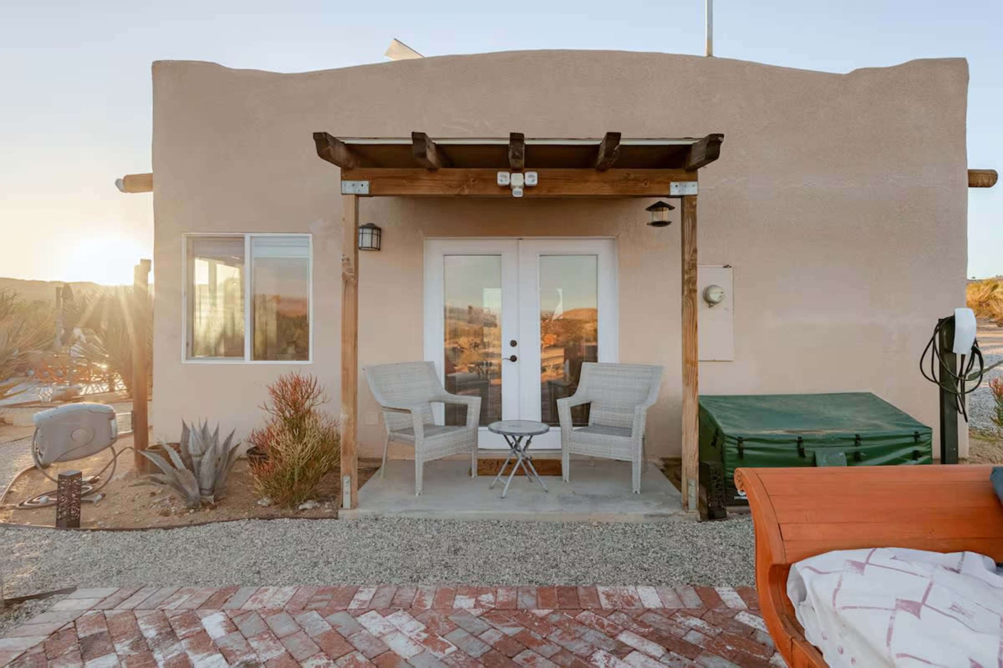 The image shows a small, stucco building with a covered porch, featuring two chairs and a table, surrounded by desert landscaping.