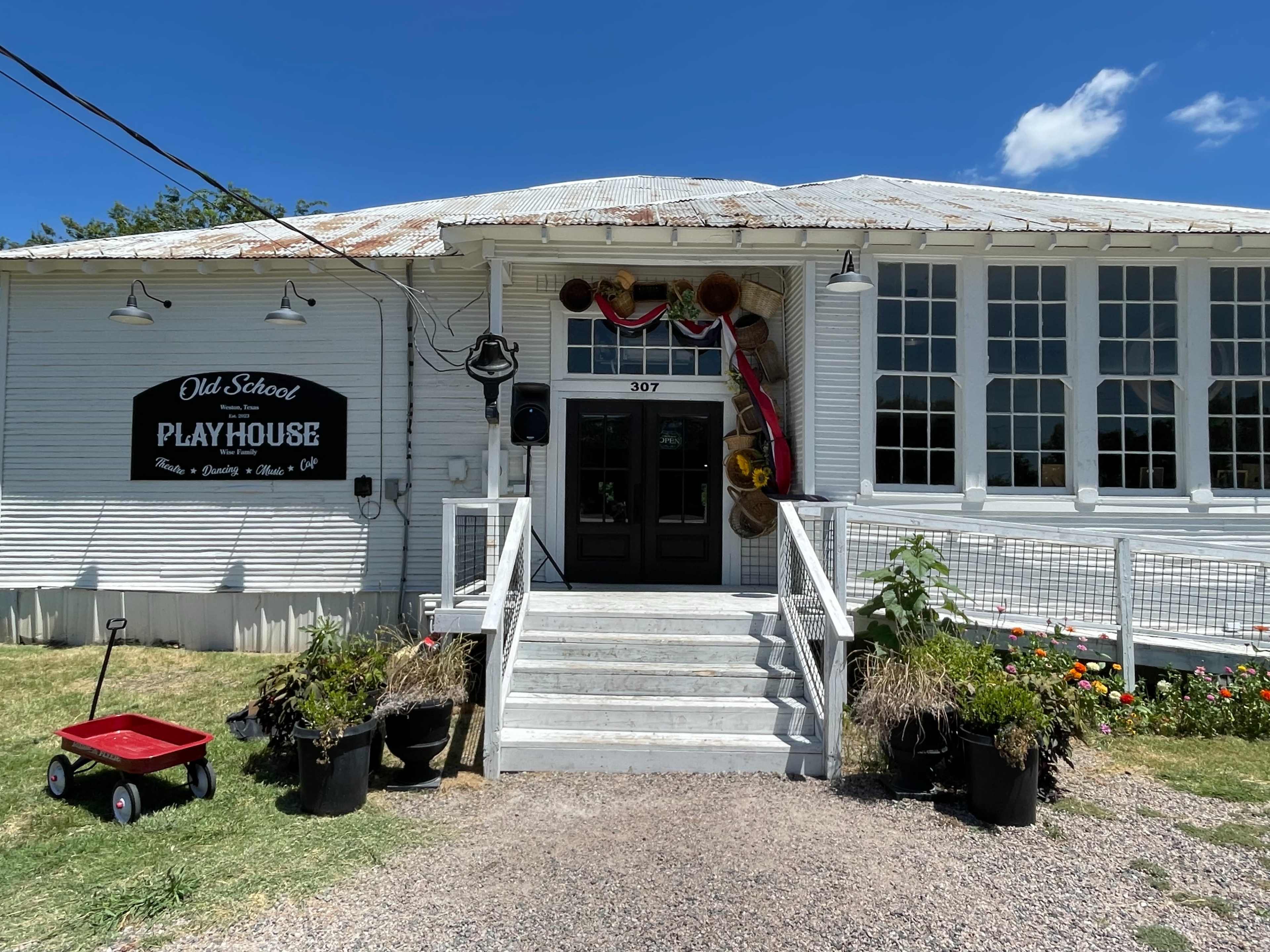 The entrance of an old playhouse, marked by a sign that reads "Old School Playhouse," with a white exterior, stairs leading up to the door, and potted plants on either side.