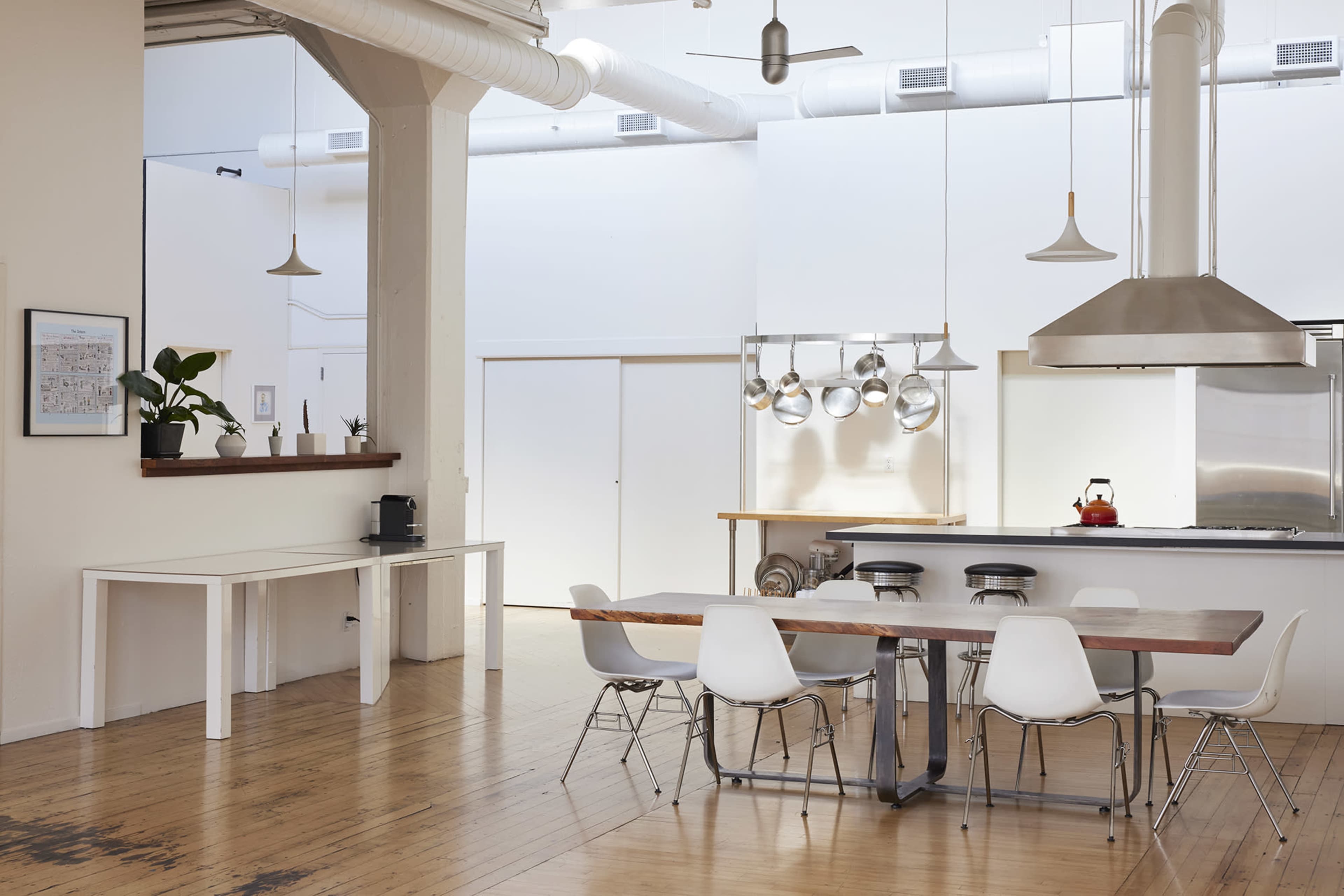 A bright and open kitchen and dining area features a large wooden table surrounded by white chairs, with stainless steel appliances and hanging pots visible in the background.