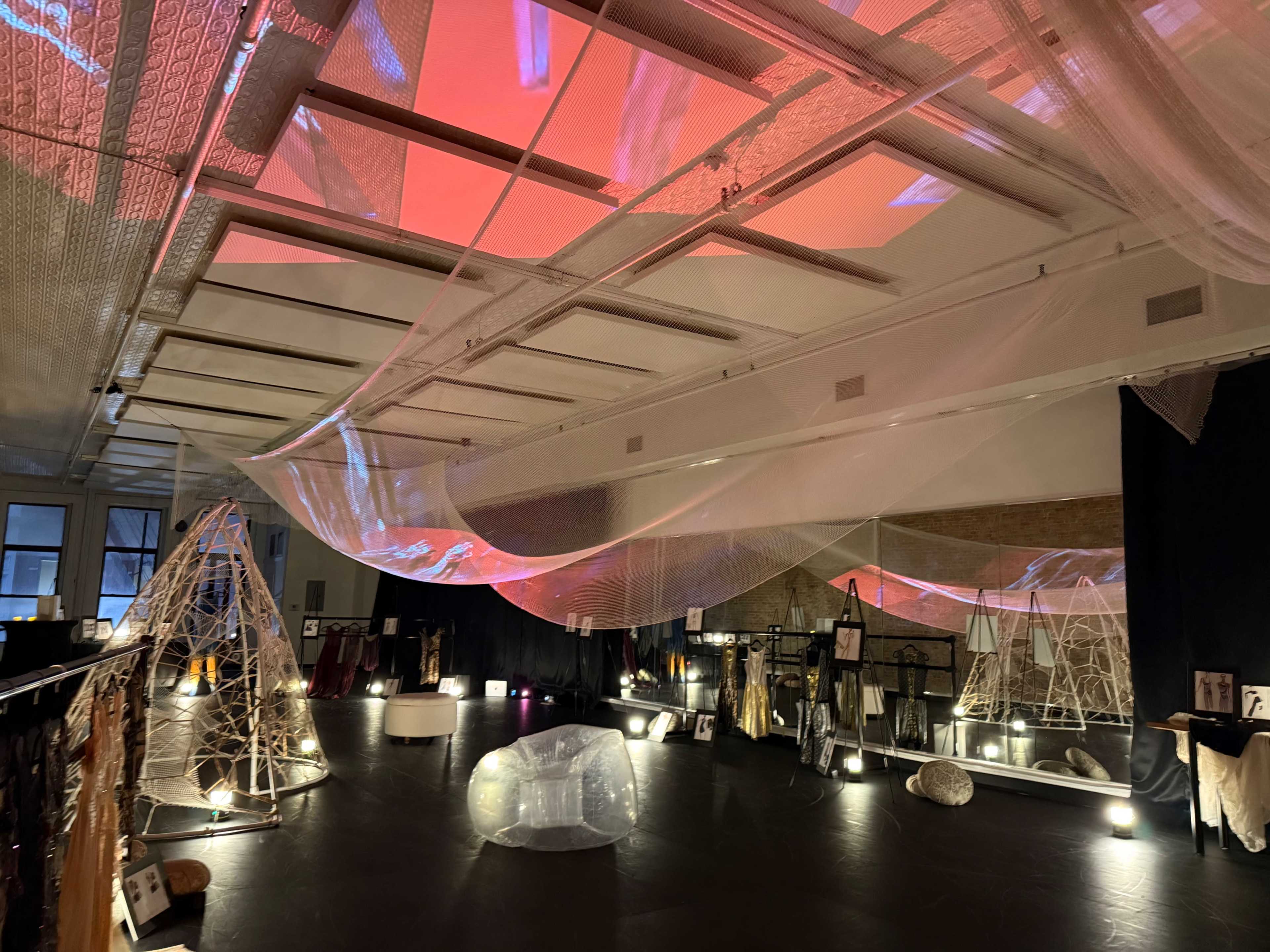 The image shows an art installation in a gallery featuring translucent fabrics suspended from the ceiling, interacting with soft lighting and various displayed sculptures.