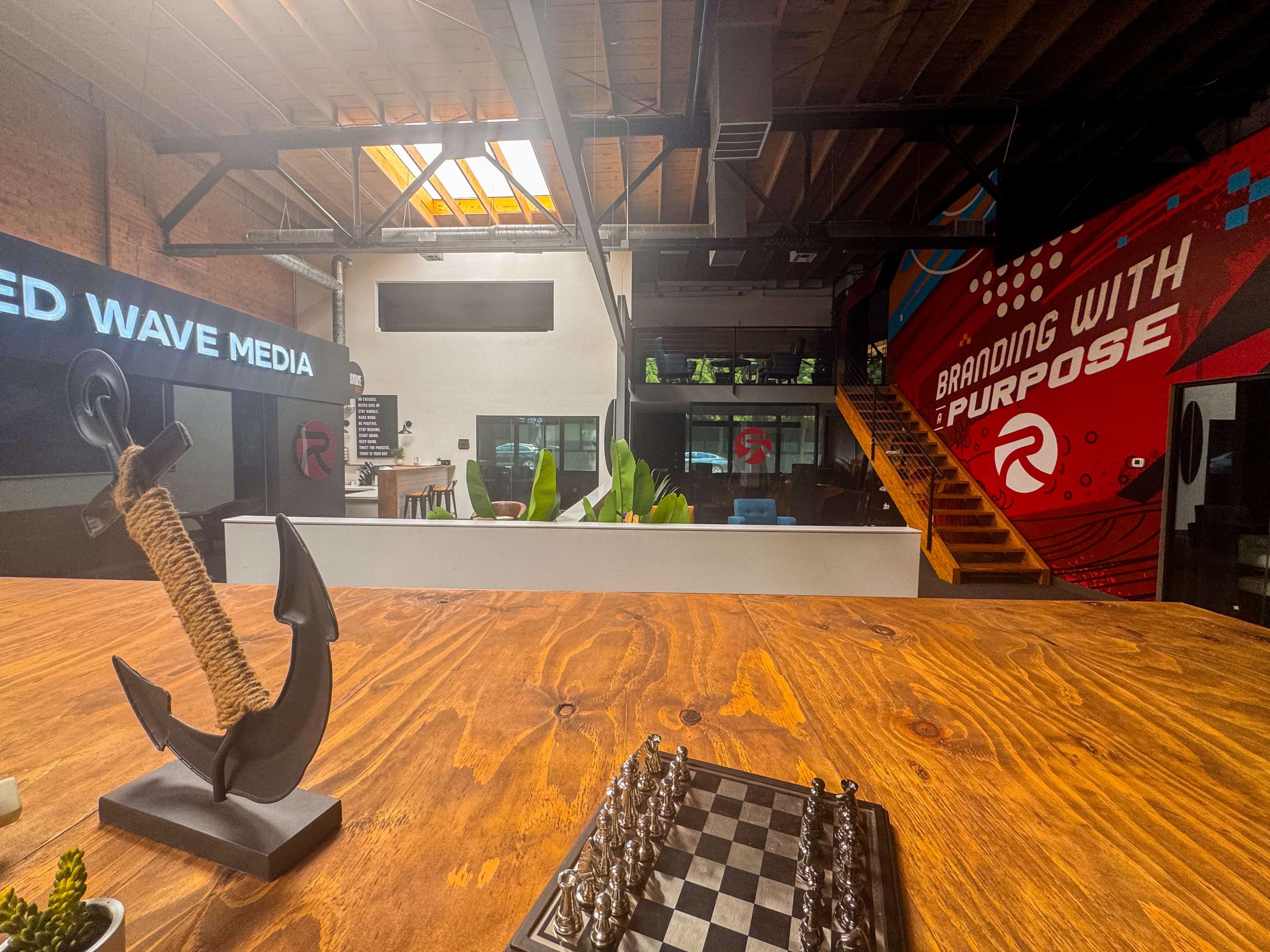 The image shows a modern office space featuring wooden tables, a chess set, large windows, and vibrant wall murals promoting branding and purpose.