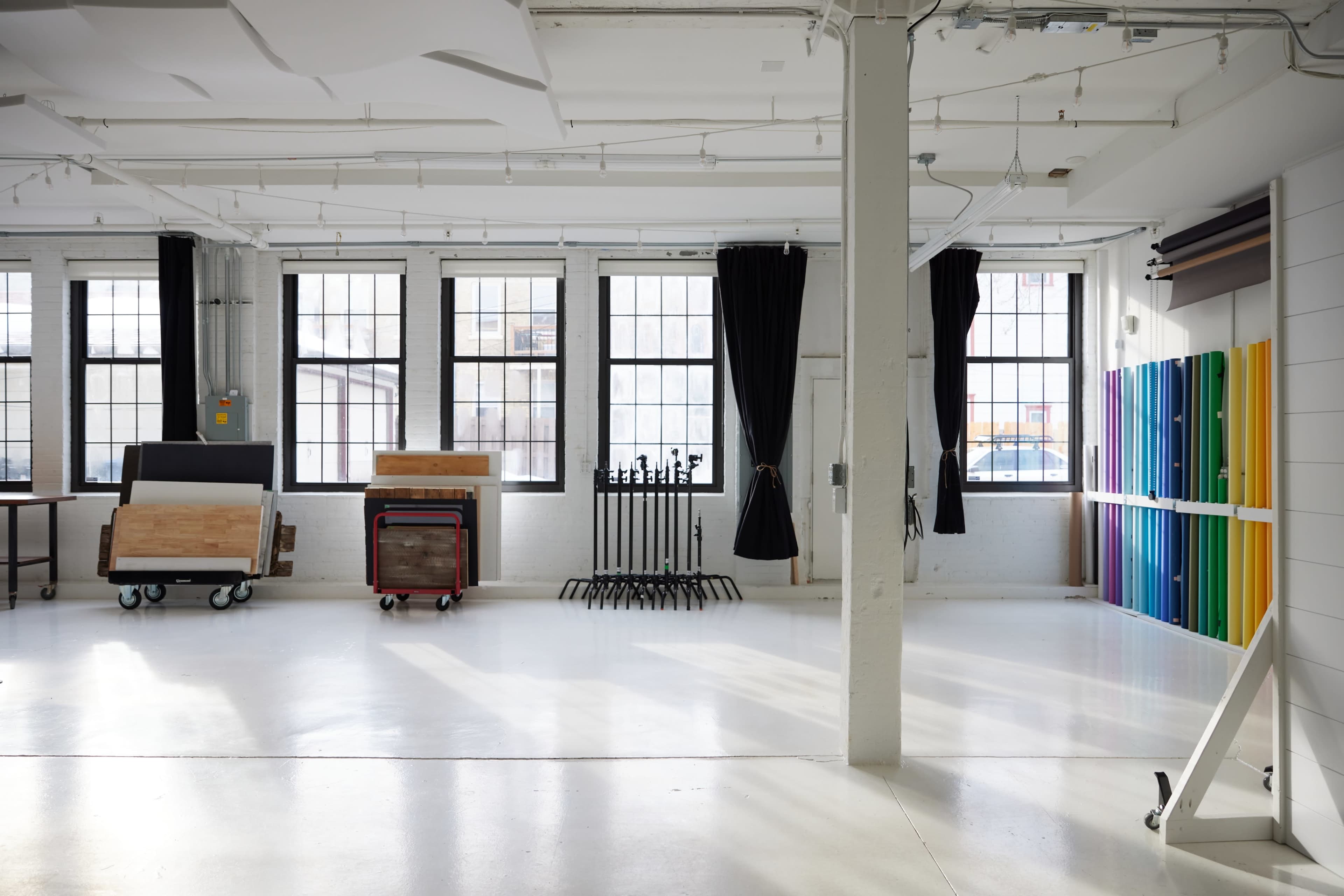 A spacious, well-lit room with large windows, wooden furniture on wheels, and colorful vertical panels arranged against one wall.