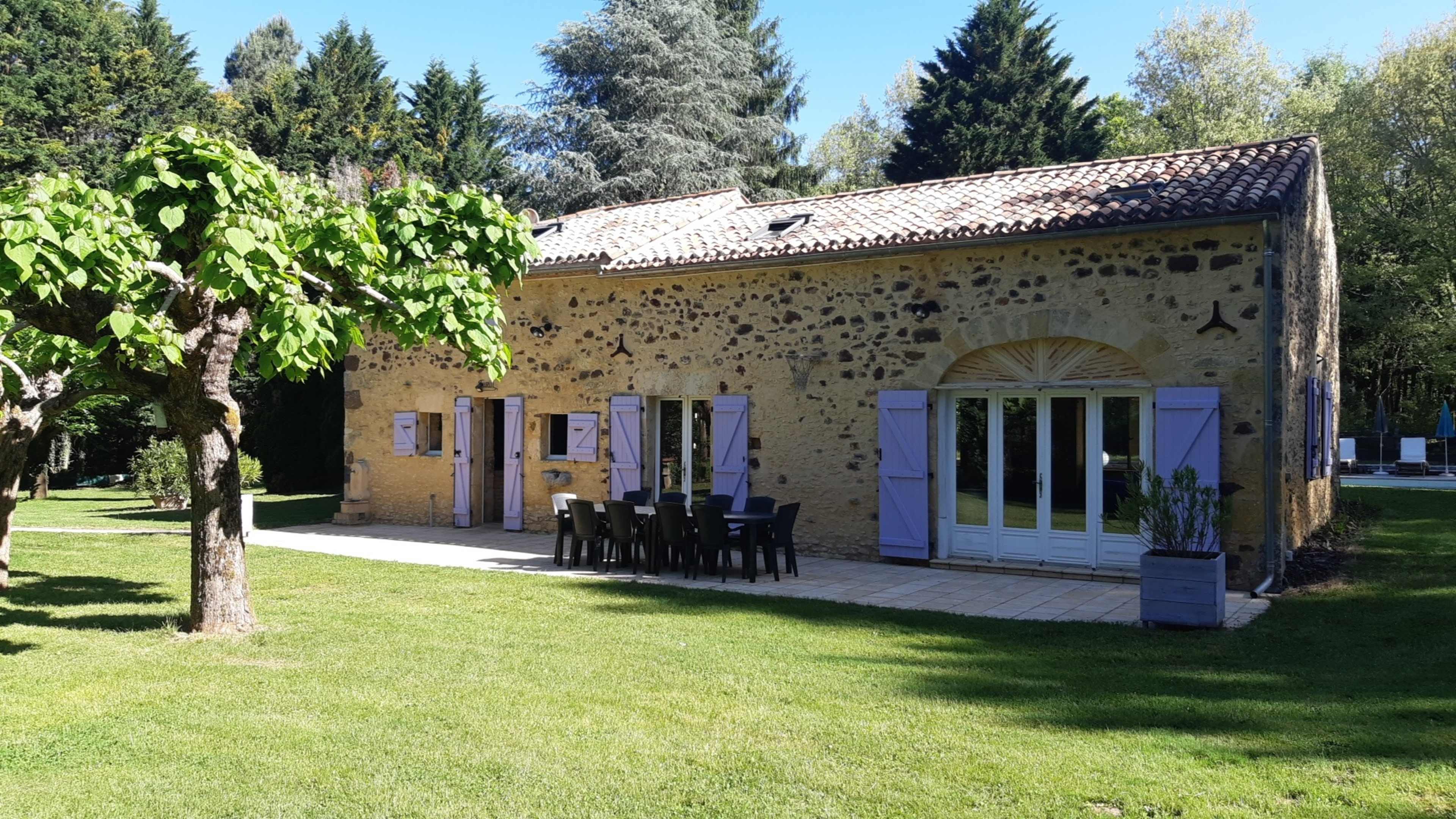 A stone house with purple shutters sits in a green yard surrounded by trees, featuring a patio with a large dining table.