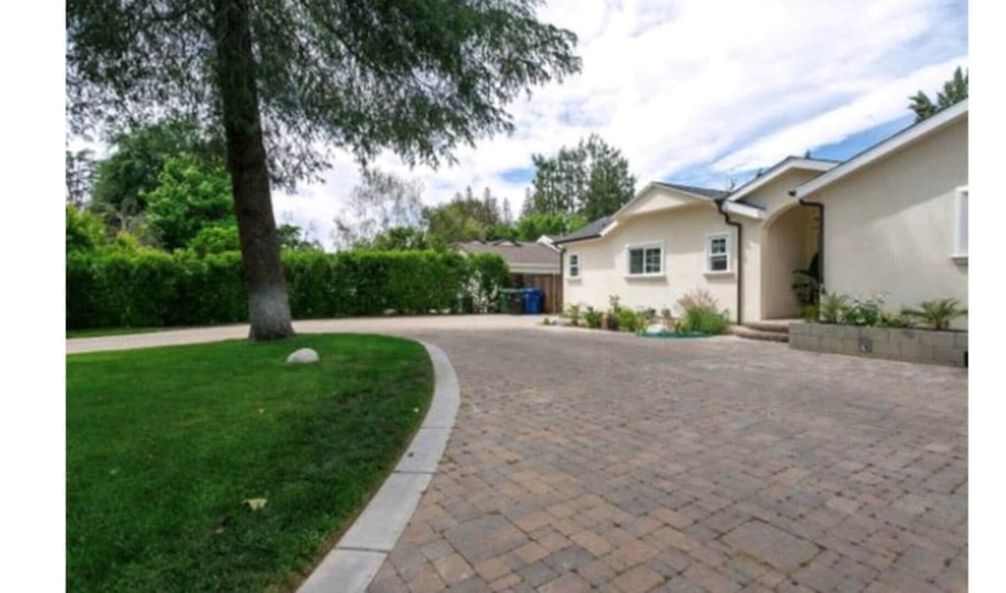 A paved driveway leads to a light-colored house surrounded by greenery and a tree on the left.