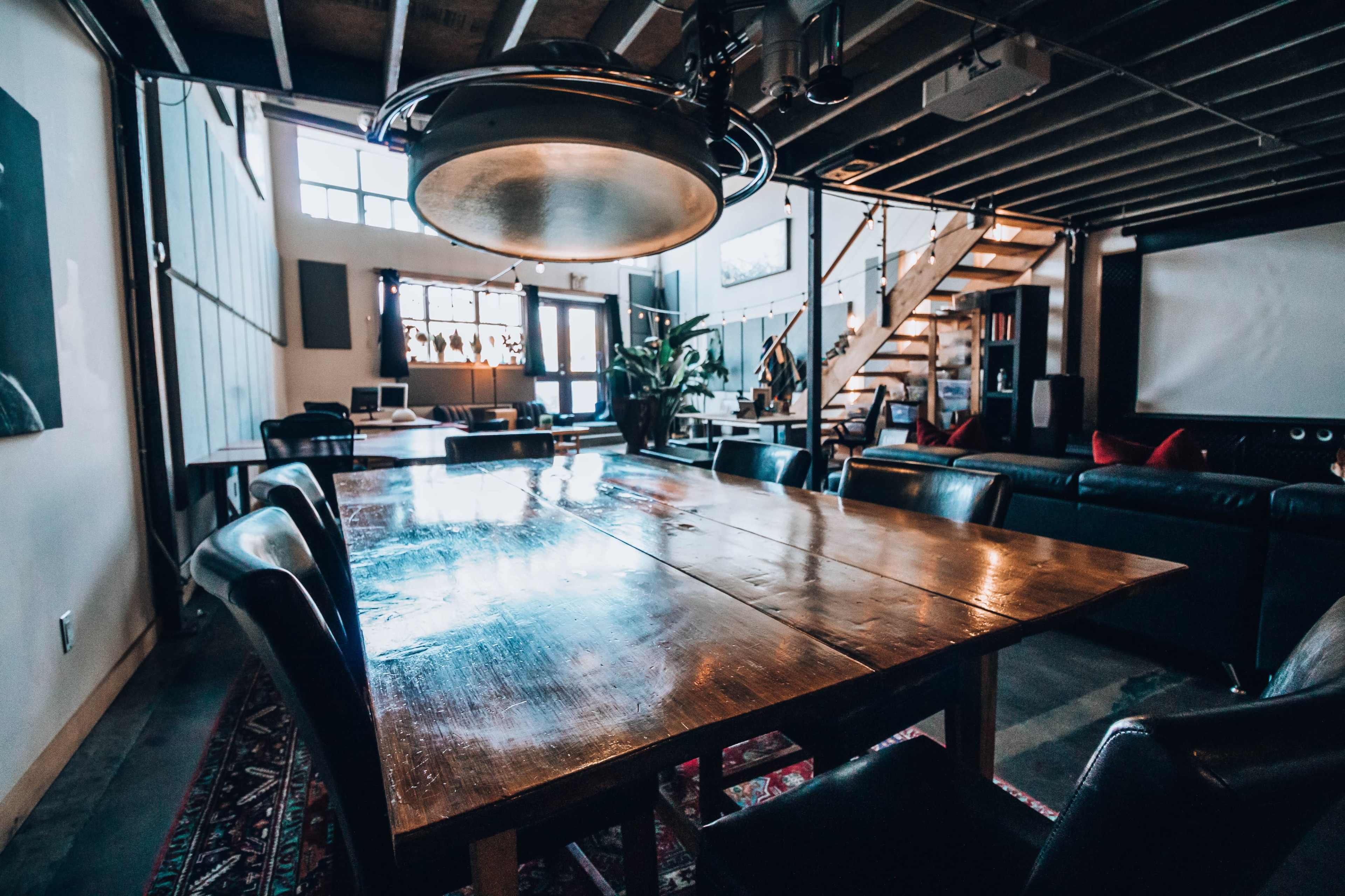 A spacious, modern living area features a large wooden dining table surrounded by black leather chairs, with a staircase and a multimedia setup in the background.