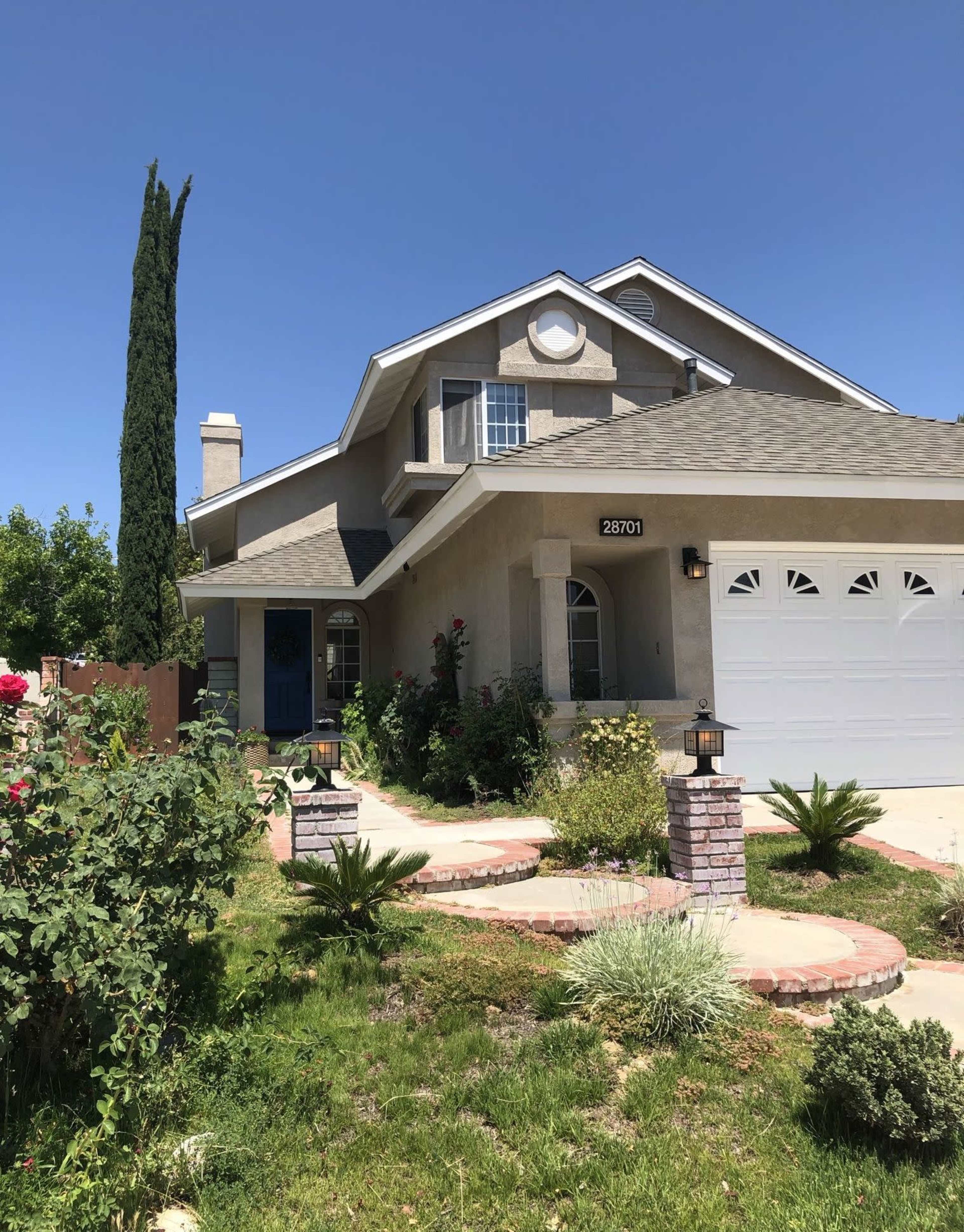 A two-story house with a gray exterior, a garage, and landscaped front yard featuring pathways, shrubs, and a tall cypress tree.