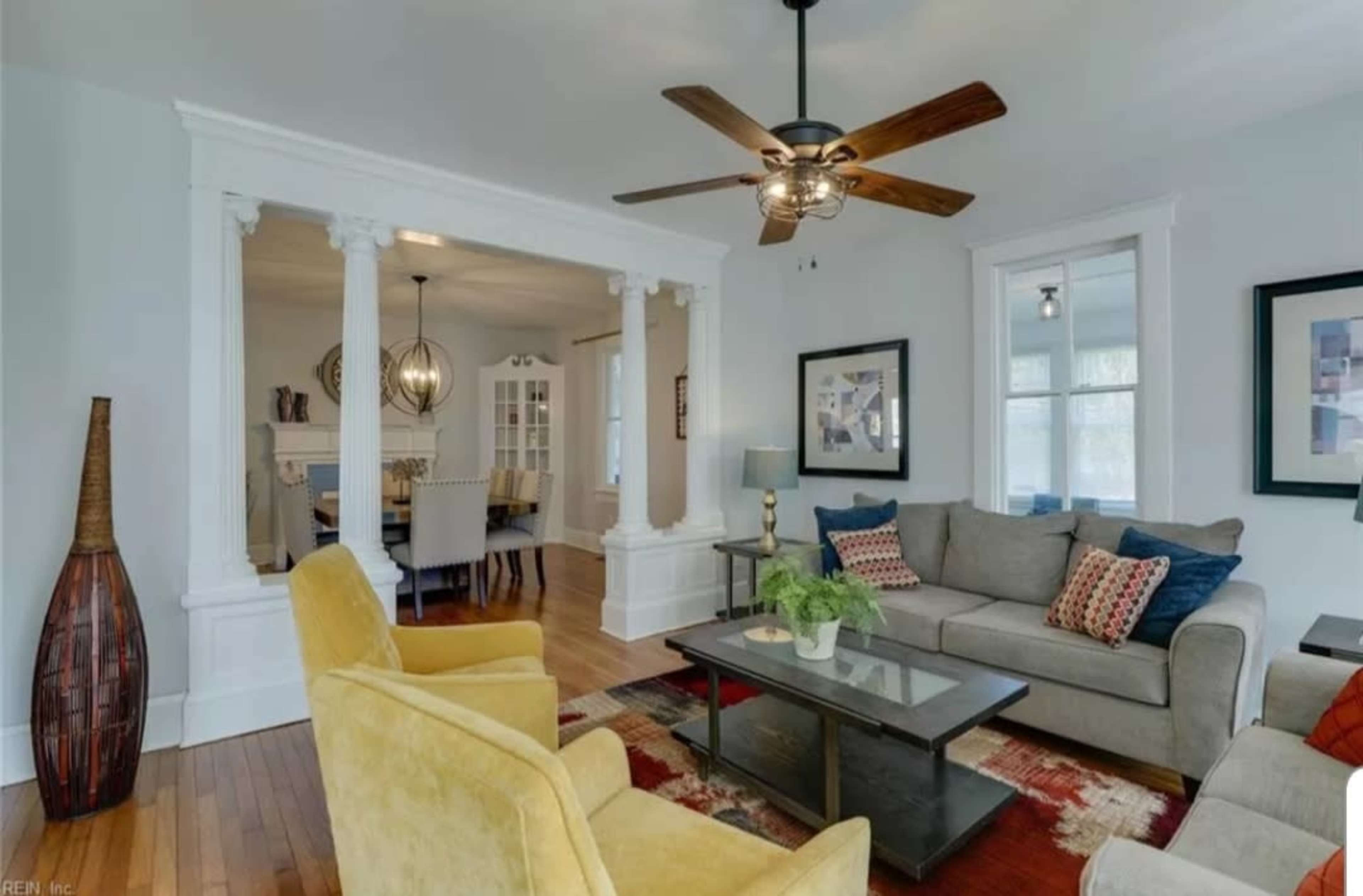 A living room with a sofa and yellow chairs, featuring a ceiling fan and a view into a dining area through a set of white columns.