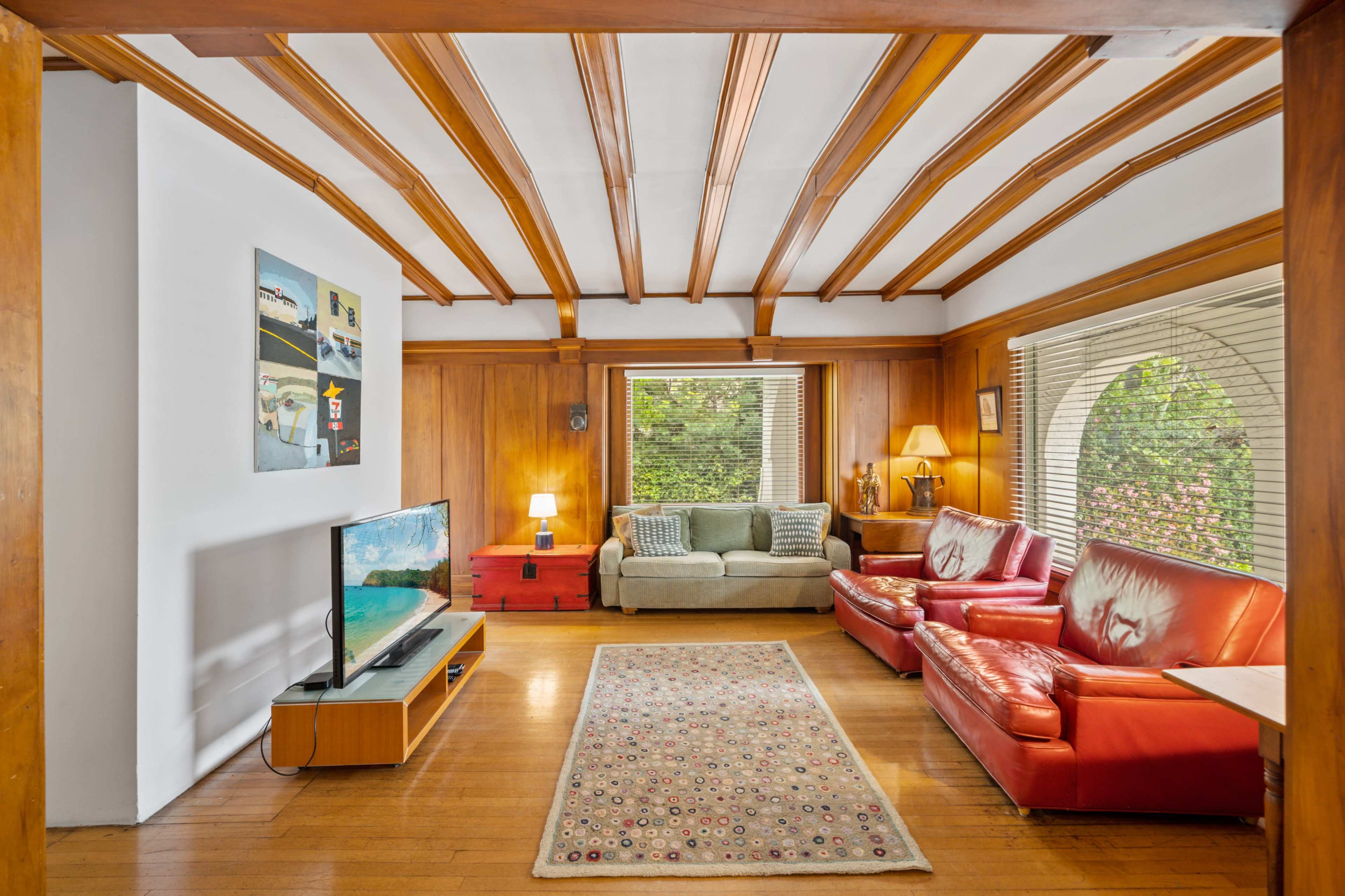 The living room features wooden beams on the ceiling, a television on a low stand, a green sofa, two red leather chairs, and a wooden chest in front of a large window.