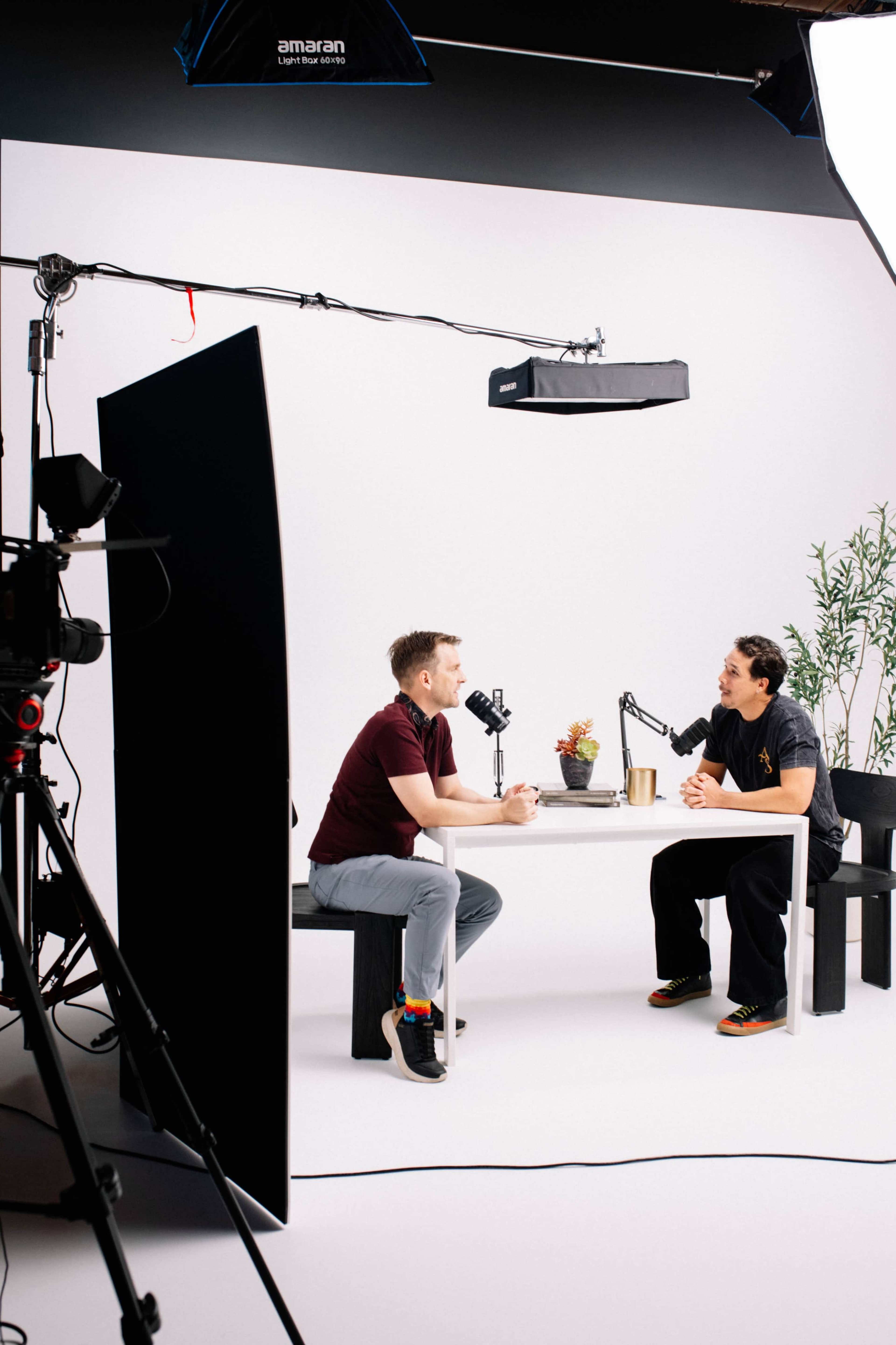Two individuals are seated at a table under studio lighting, engaged in a conversation with microphones positioned in front of them.