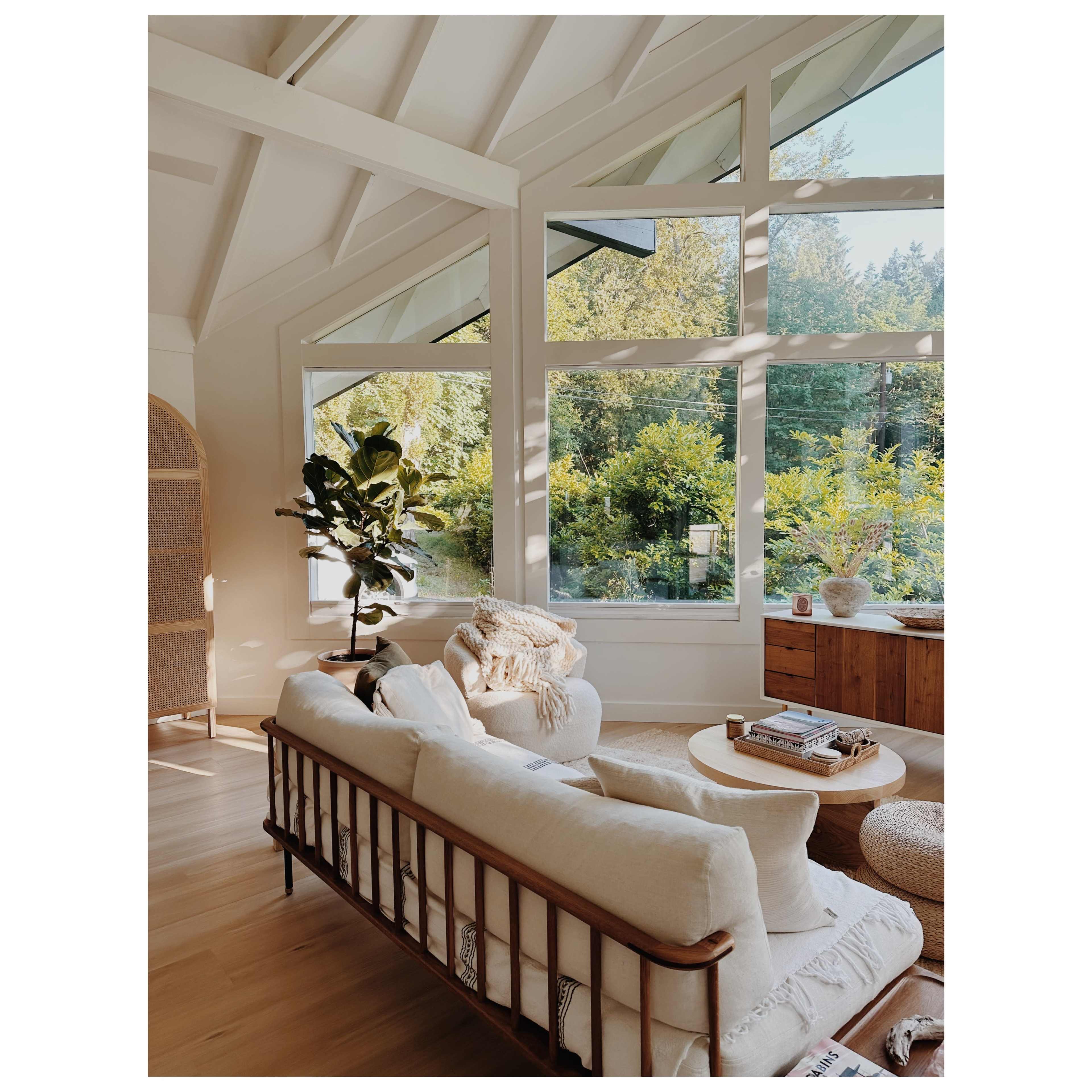 A bright living room features a white sofa, a round coffee table, and large windows overlooking a lush garden.