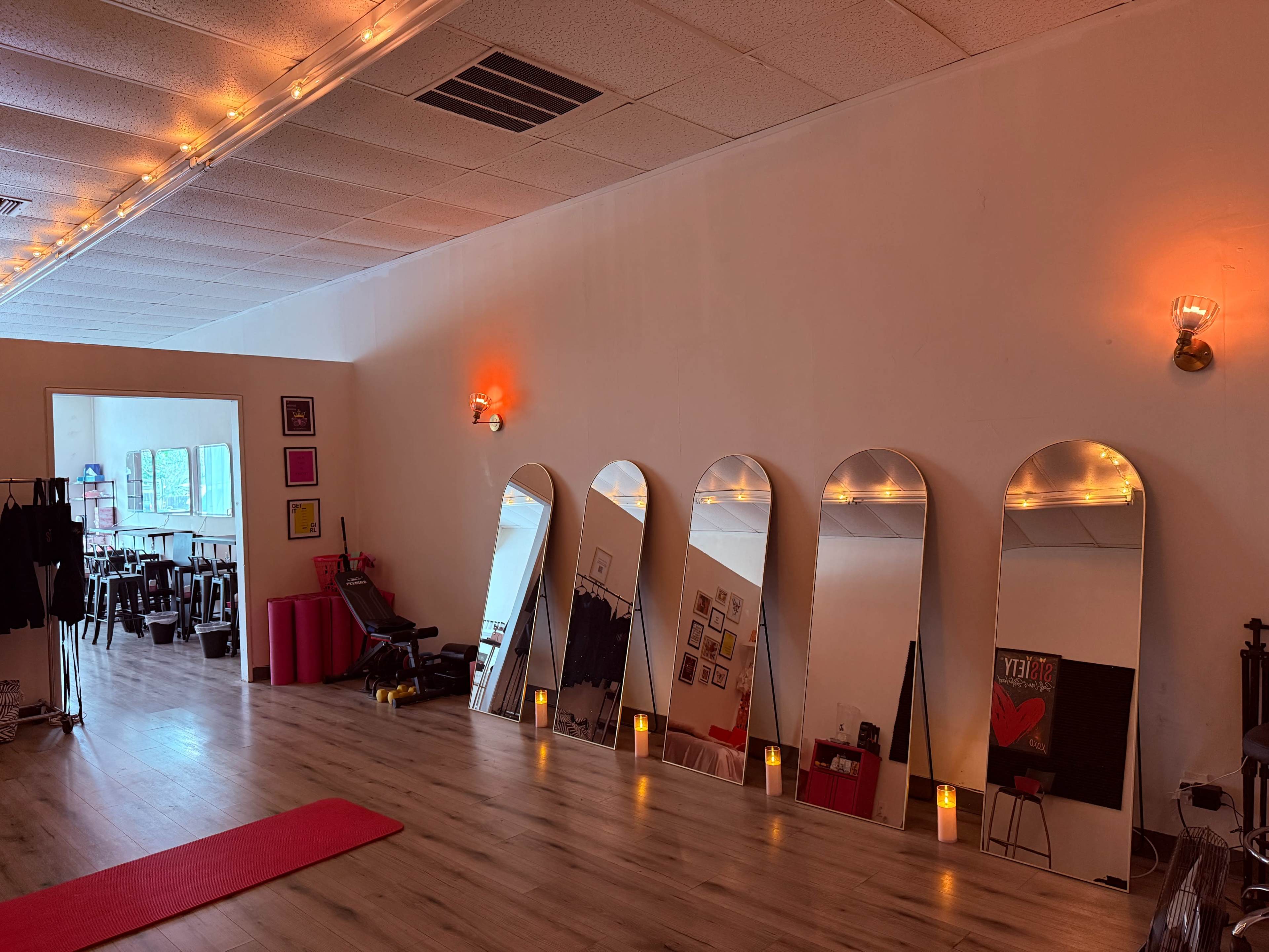 The image shows an exercise or yoga studio featuring several large mirrors along one wall, with soft lighting and a red mat on the wooden floor.