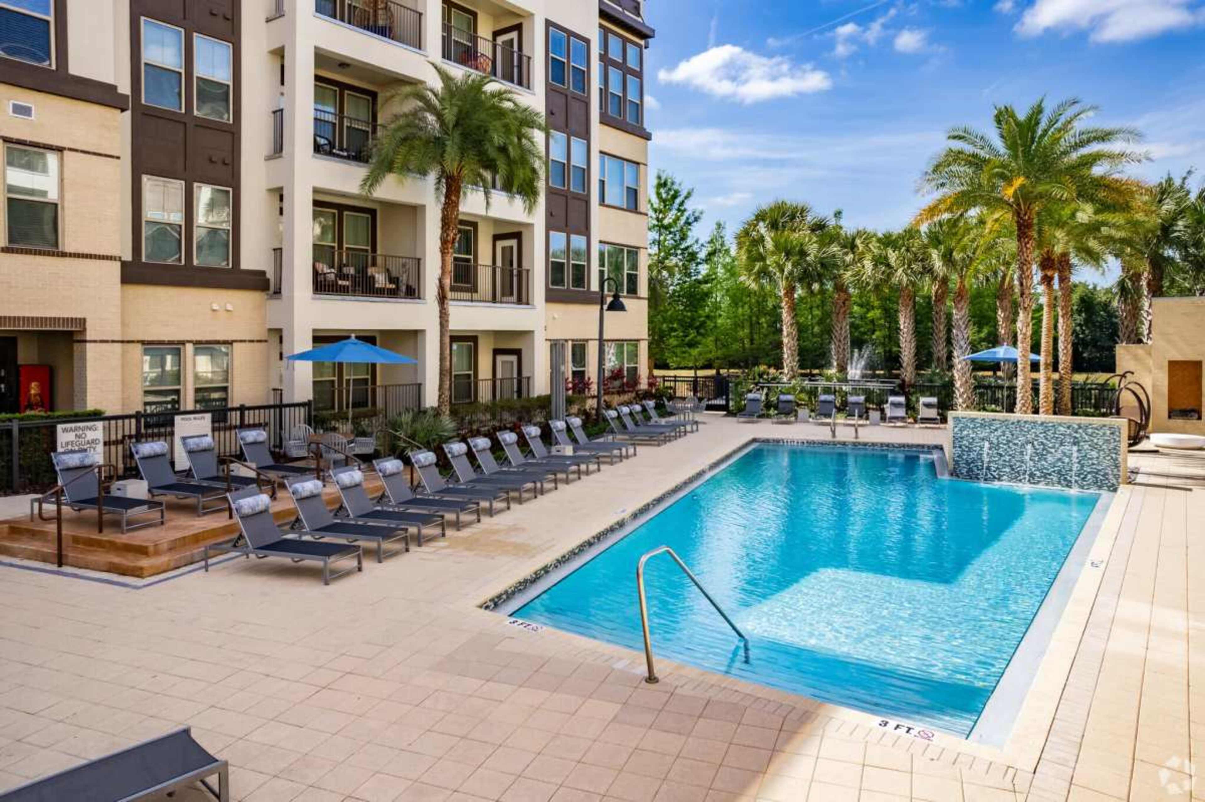 The image shows a swimming pool surrounded by lounge chairs and palm trees, adjacent to an apartment building.